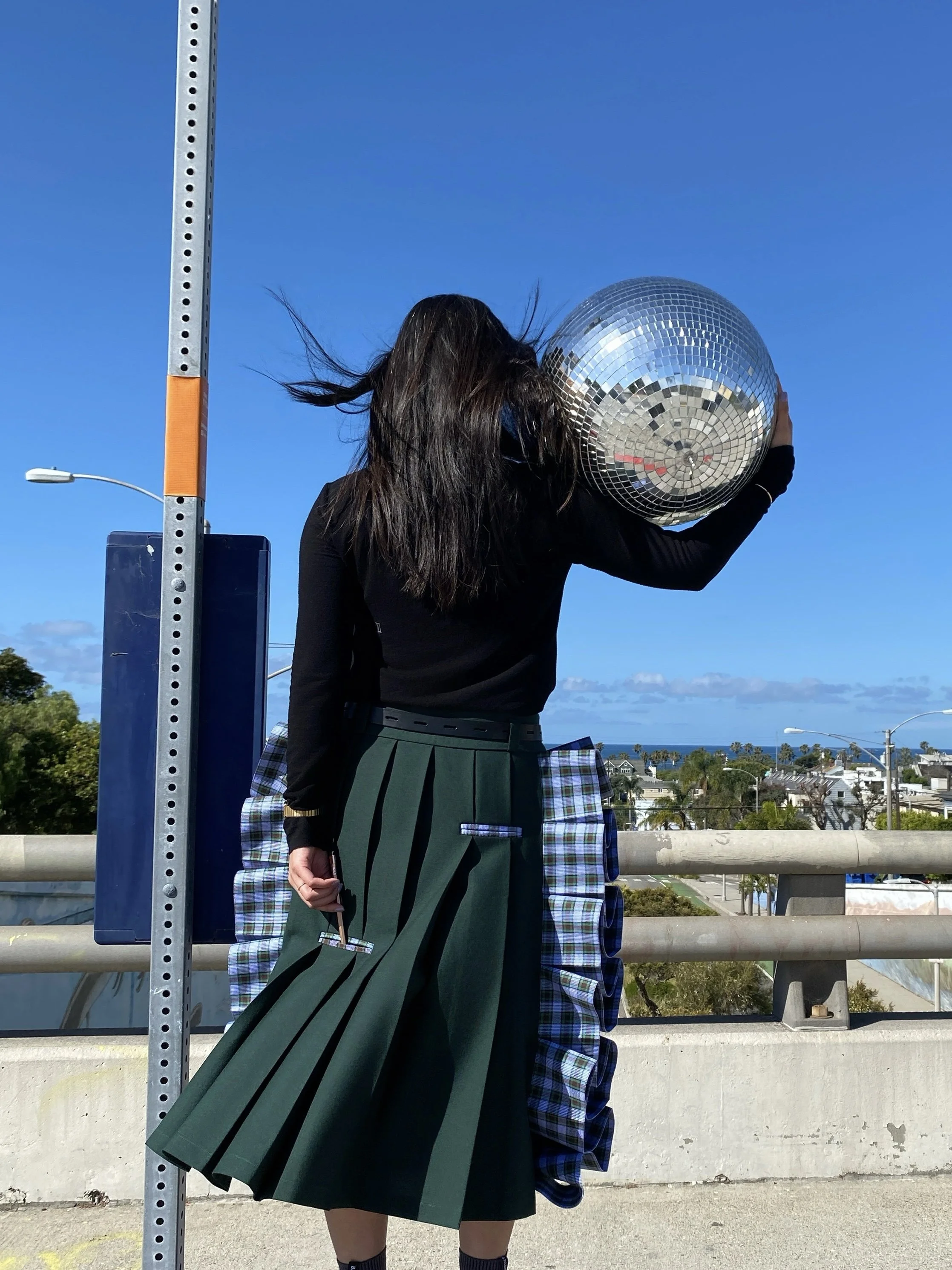 A woman standing outdoors on a sunny day, holding a large disco ball near her ear, with her face obscured by the ball. She has long dark hair, is wearing a black top, a green pleated skirt, and plaid fabric hanging from the skirt. The background show