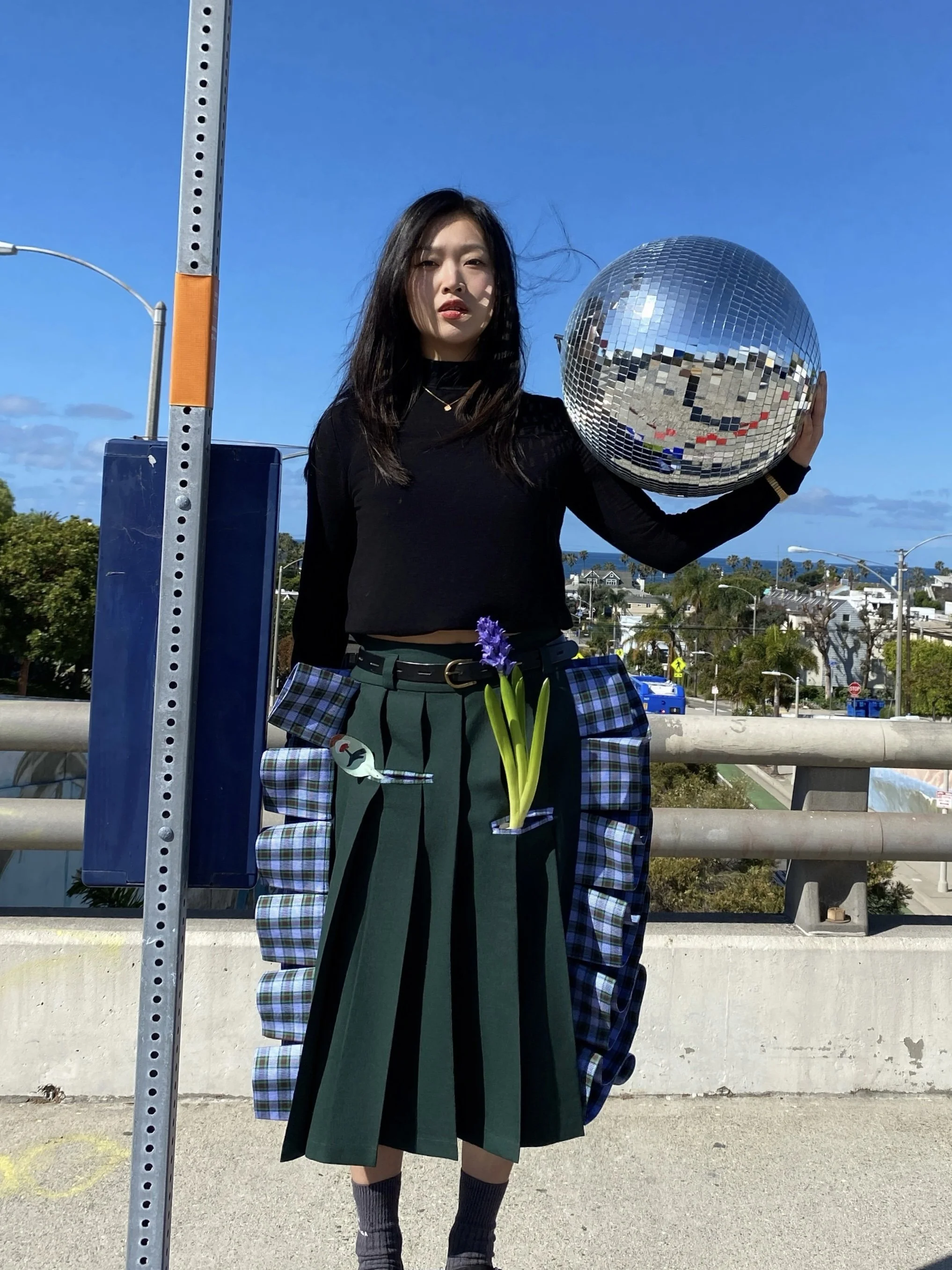 A woman in pleated skirt holding a reflective disco ball in an outdoor setting with blue sky in the background and ocean in the distance.