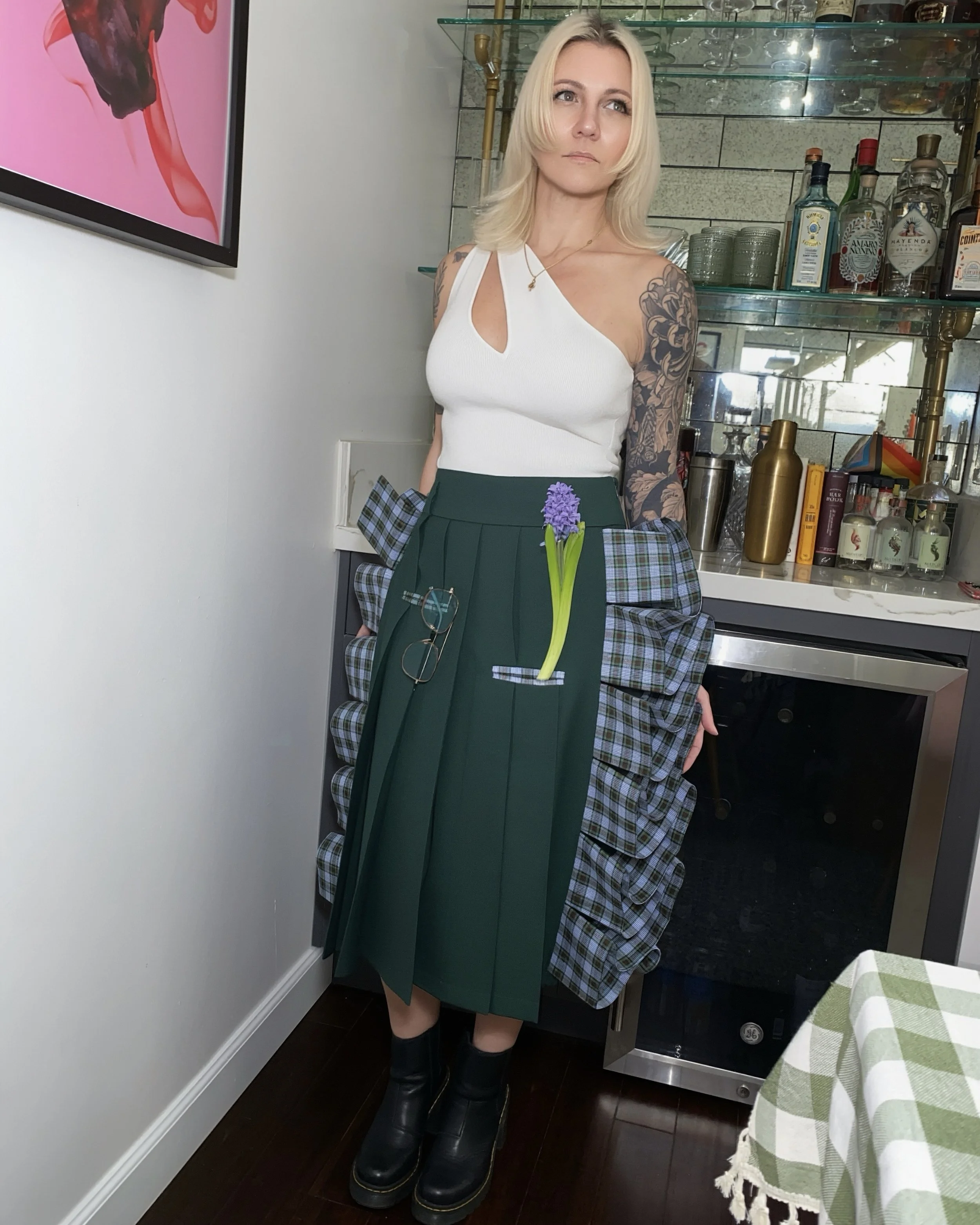 A woman with blonde hair and tattoos on her arms standing in a bar or restaurant setting. She is wearing a white asymmetrical top and a green pleated skirt with pockets holding a purple hyacinth flower. She has black boots and glasses hanging from he