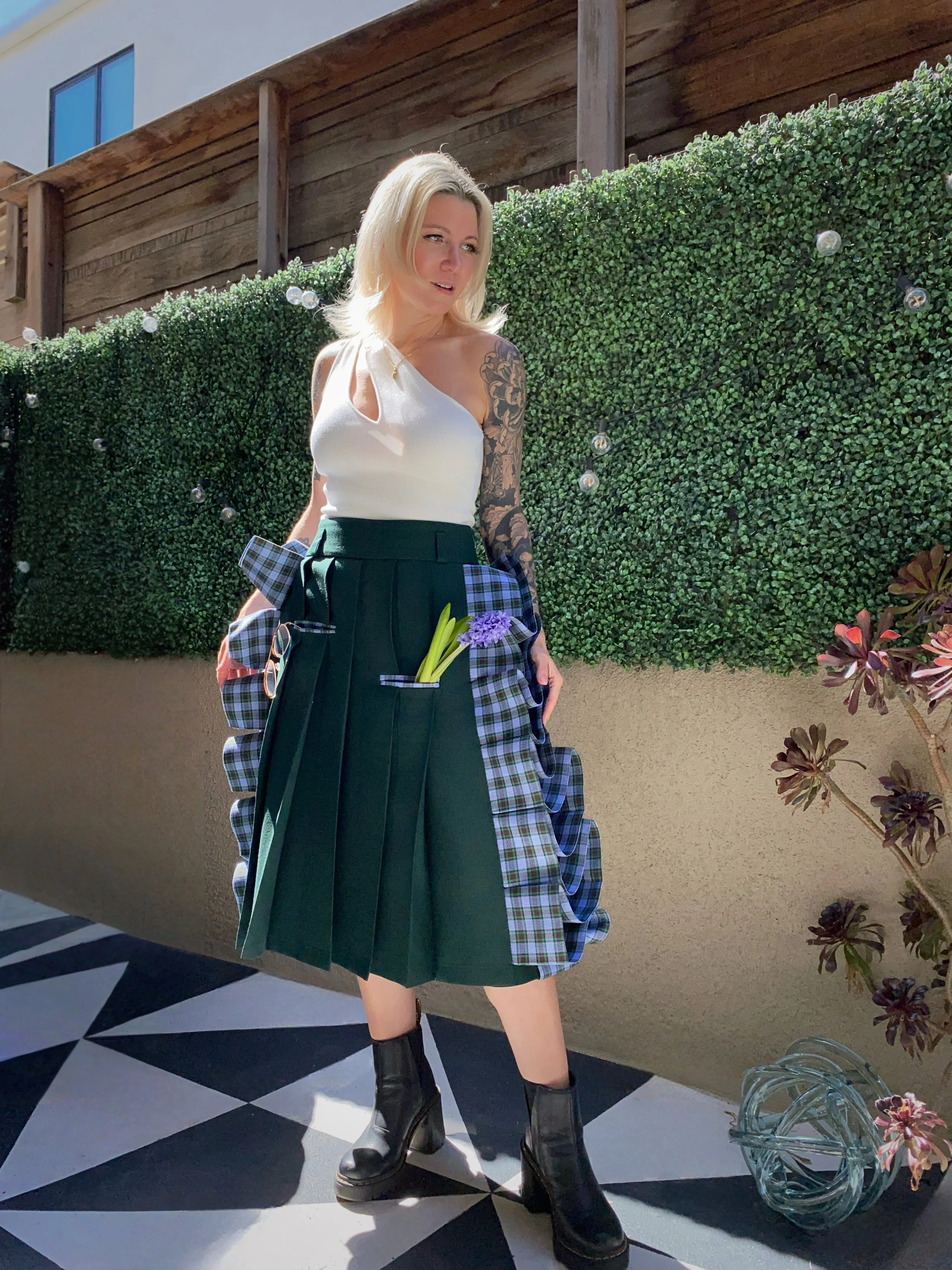 A woman with blonde hair wearing a white sleeveless top, a green pleated skirt with plaid fabric accents, and black ankle boots, standing outdoors in front of a green hedge with string lights and a wooden fence.