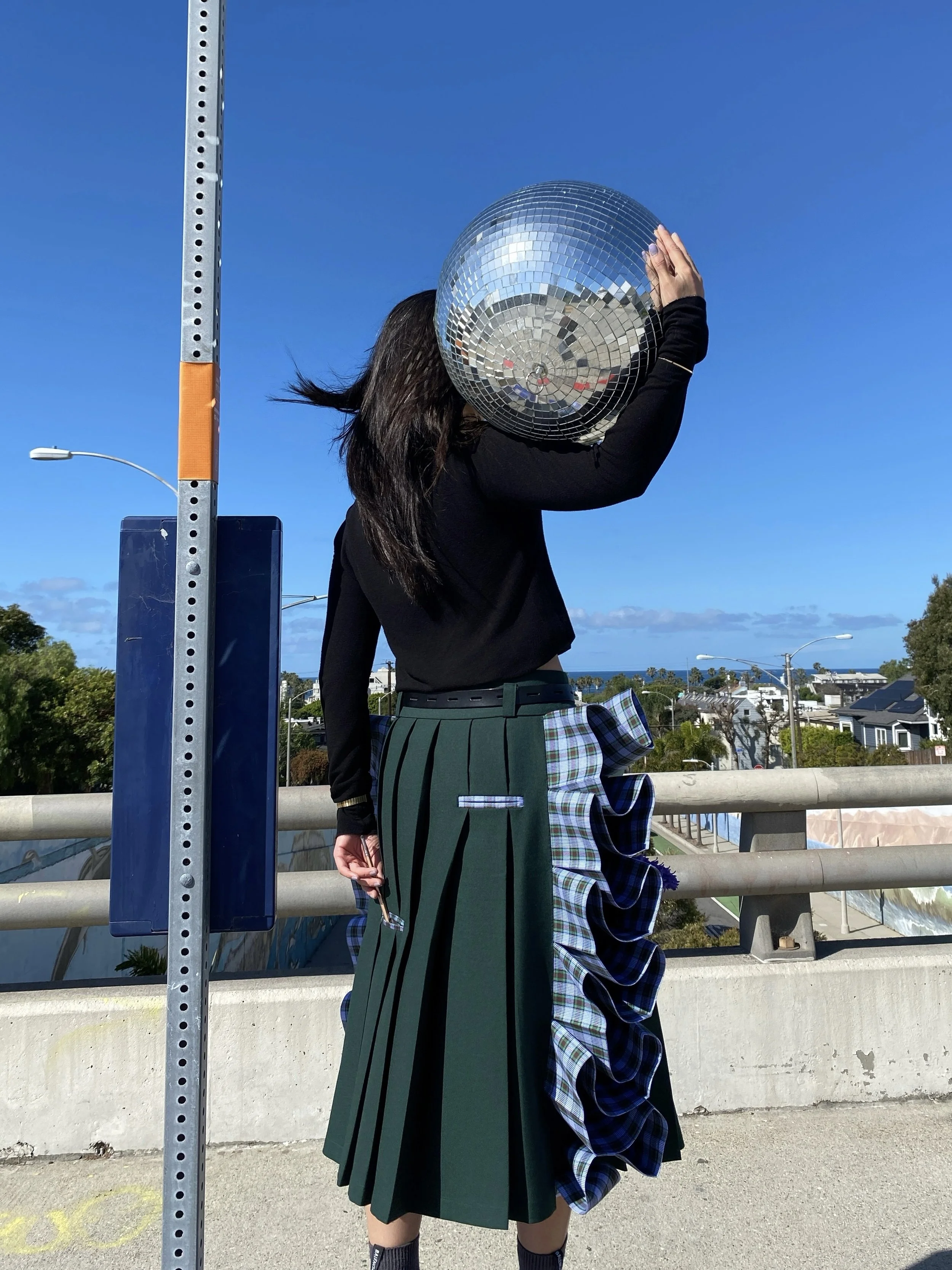 A person standing outdoors on a bridge holding a disco ball in front of their face. They are wearing a black top and a green skirt with a side ruffle with a plaid pattern. The background features a clear blue sky and some trees and buildings in the d