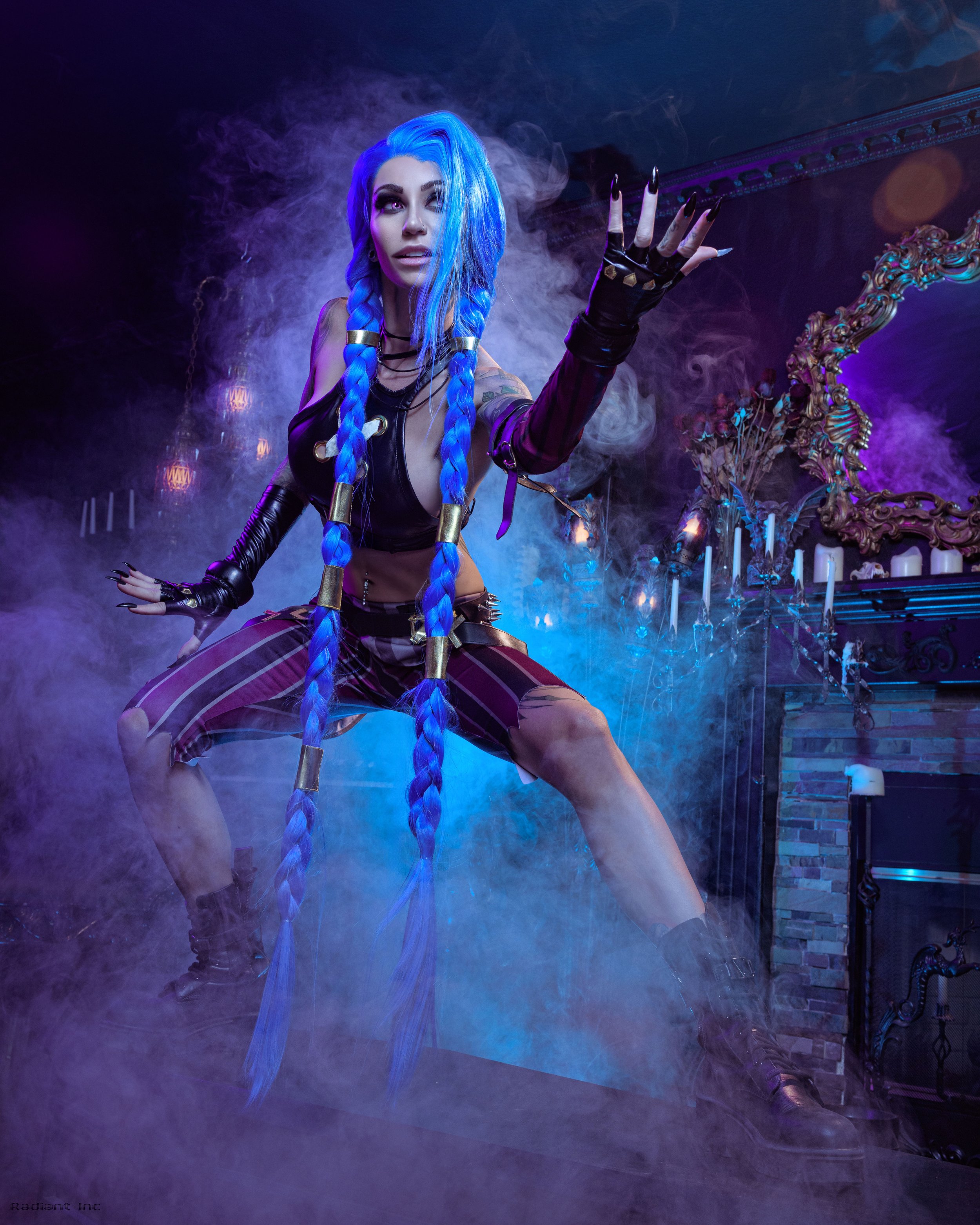 A woman with long blue braided hair and dark makeup poses in a dimly lit, foggy room with purple and blue lighting. She wears a black outfit with striped pants, black gloves, and is reaching out with her hand.