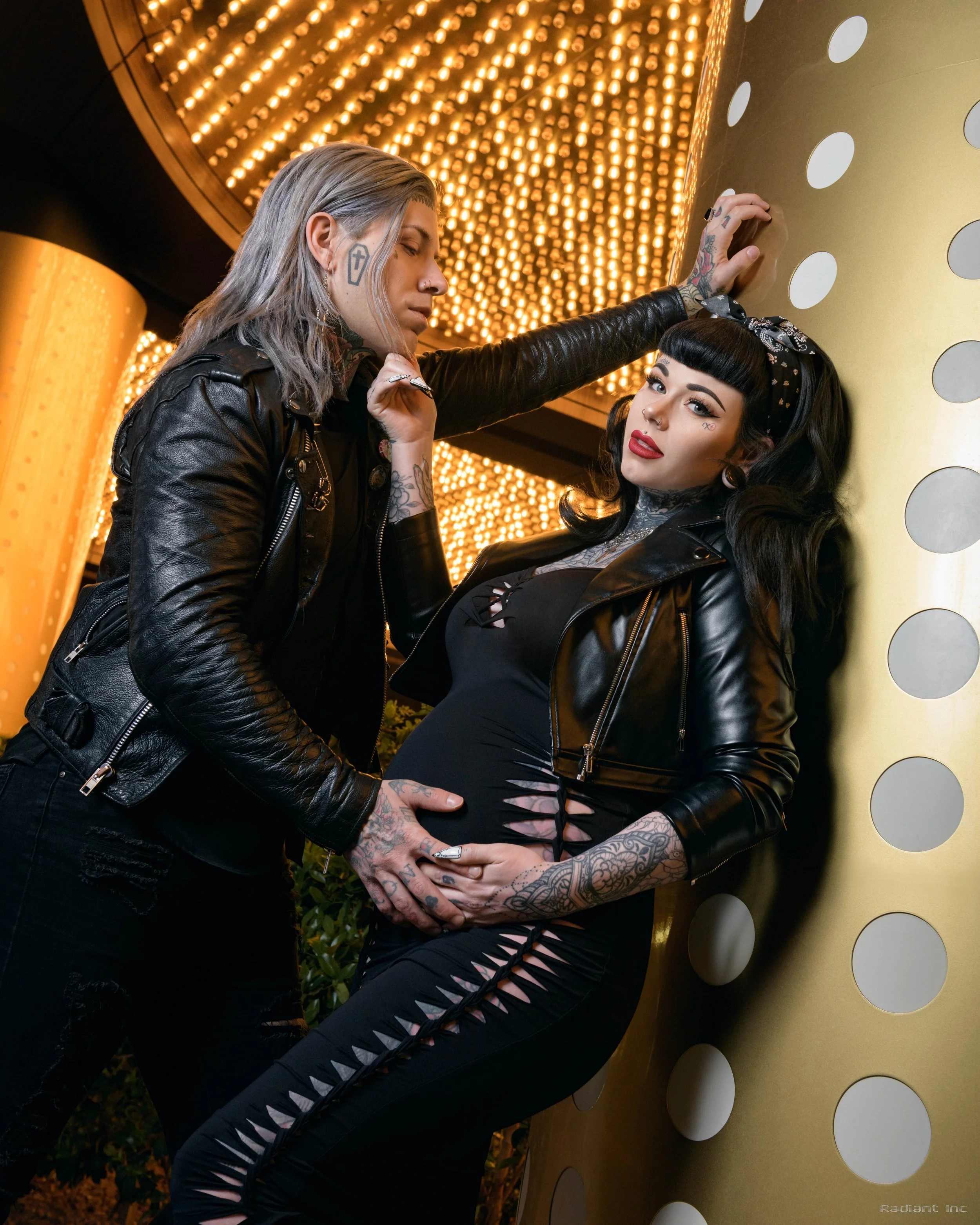 A man and woman with tattoos and dark clothing pose together indoors against a shiny gold and dotted wall, with warm lights overhead.