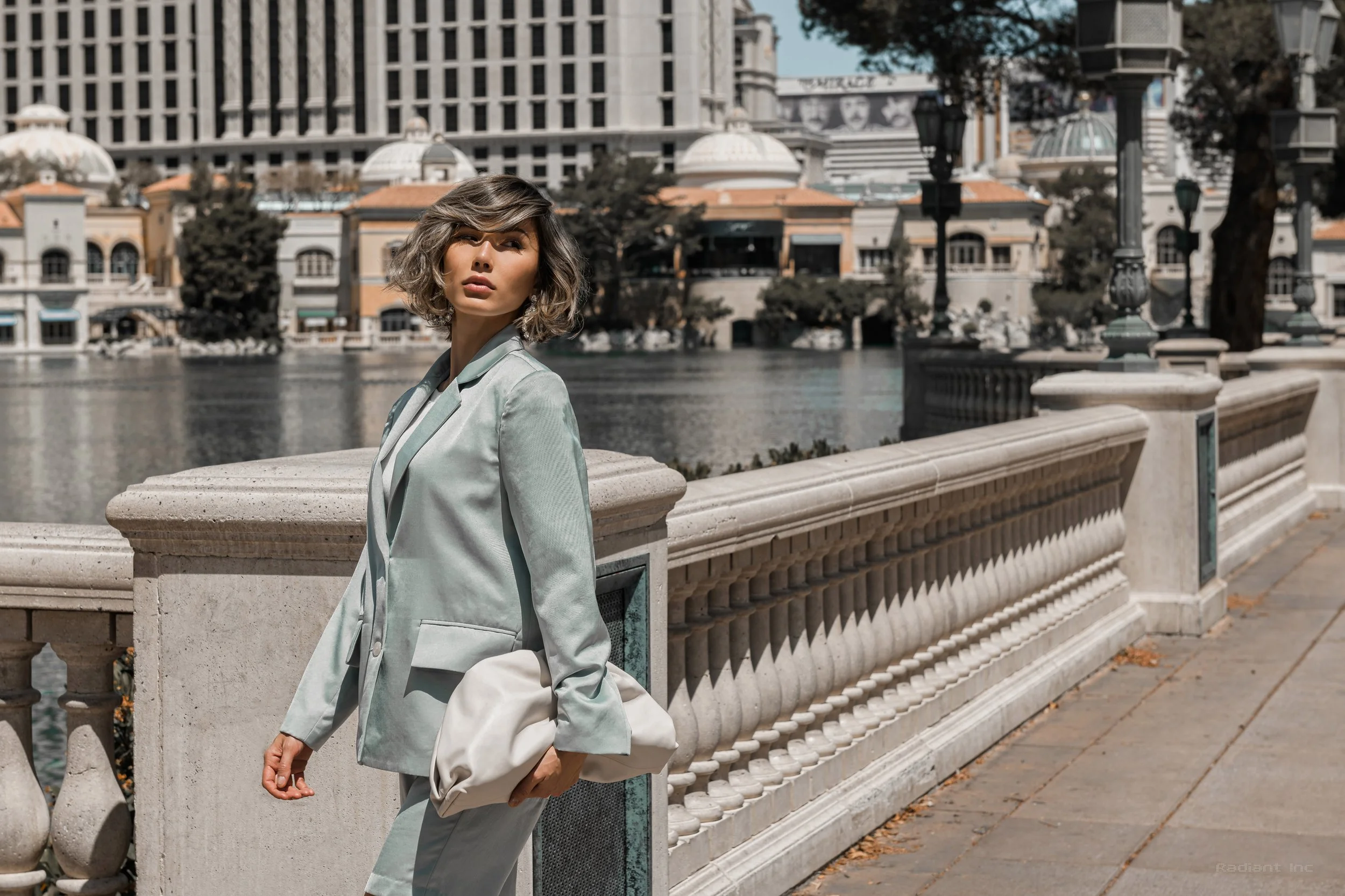 A woman in a light gray suit holding a white bag, standing on a decorative stone bridge by a river in an urban area with modern buildings and trees in the background.