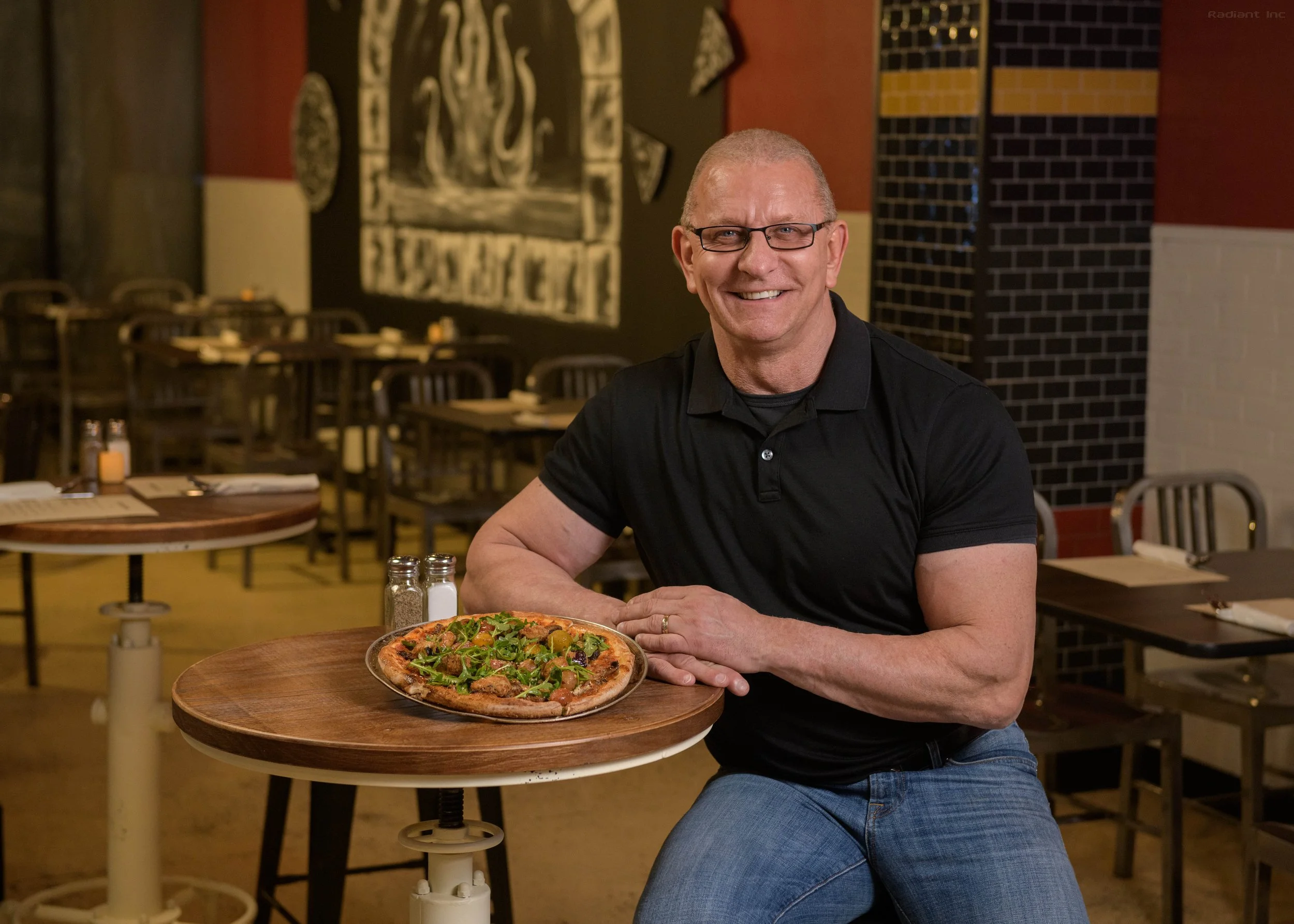 A smiling man wearing glasses and a black polo shirt, sitting at a round wooden table in a restaurant, with a freshly baked pizza topped with greens and meats in front of him.