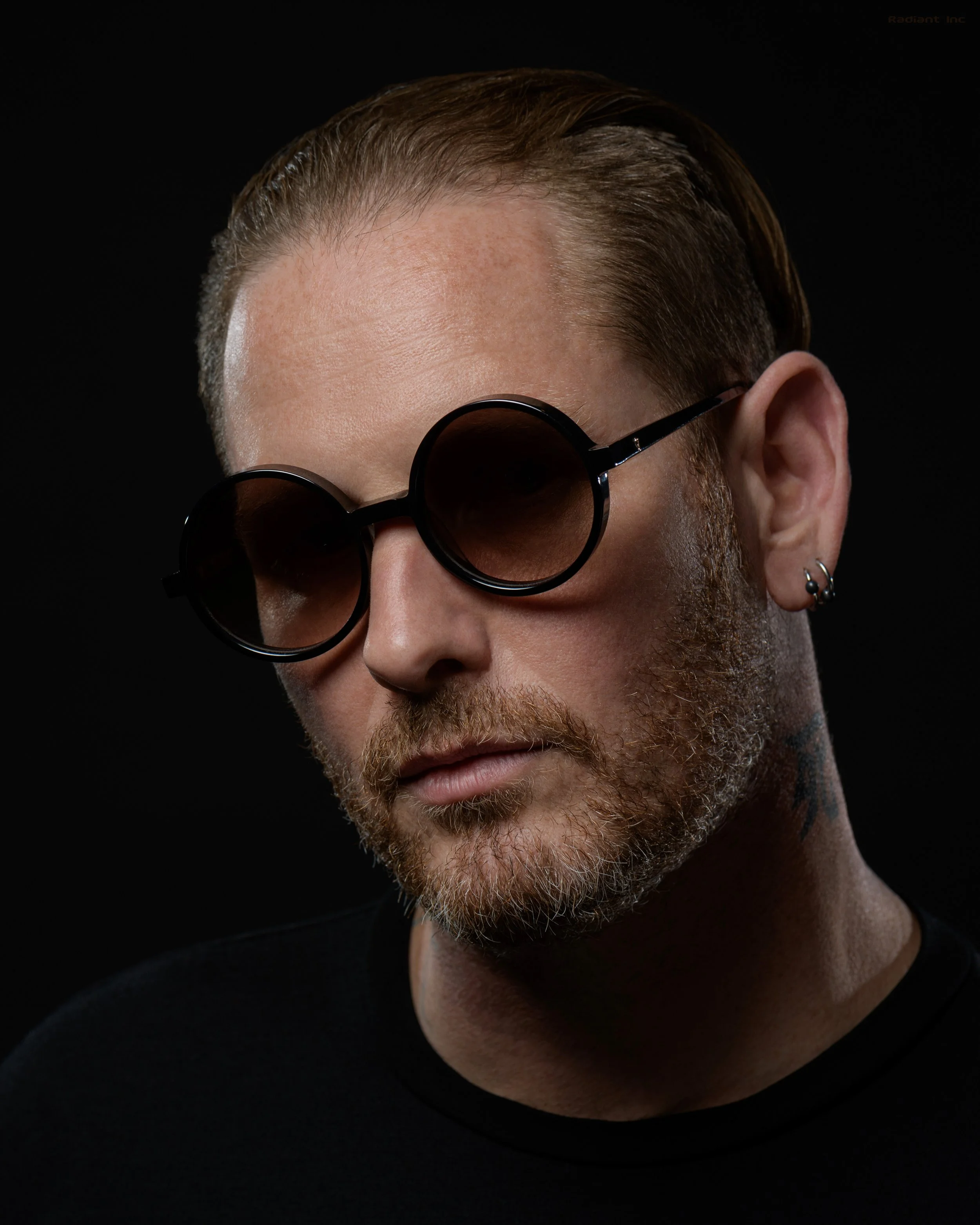 A man with short hair, a beard, and multiple earrings wearing round sunglasses and a black shirt against a black background.