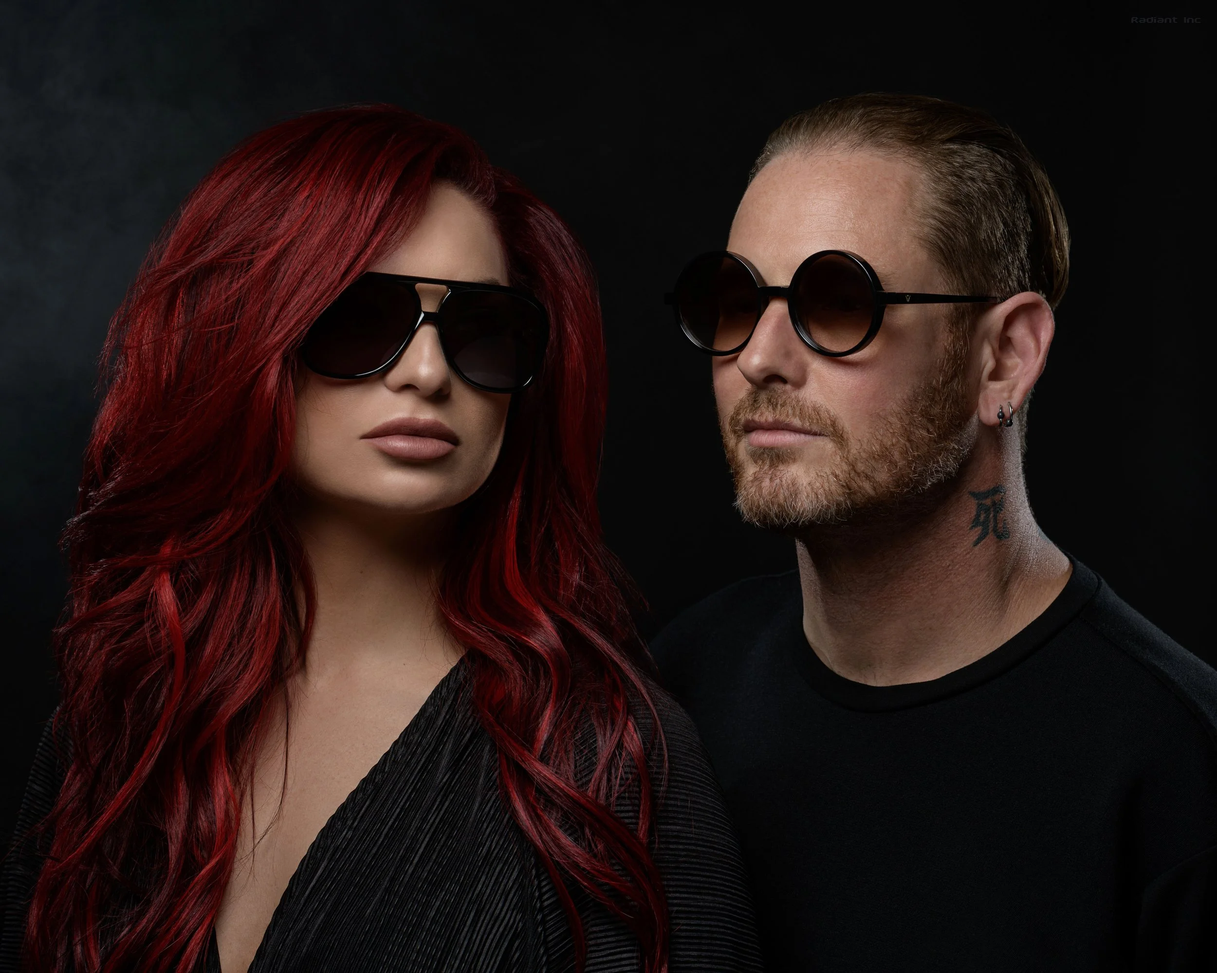 A woman with long red hair wearing large black sunglasses and a black textured top, standing next to a man with short hair, a beard, sunglasses, and earrings, against a dark background.