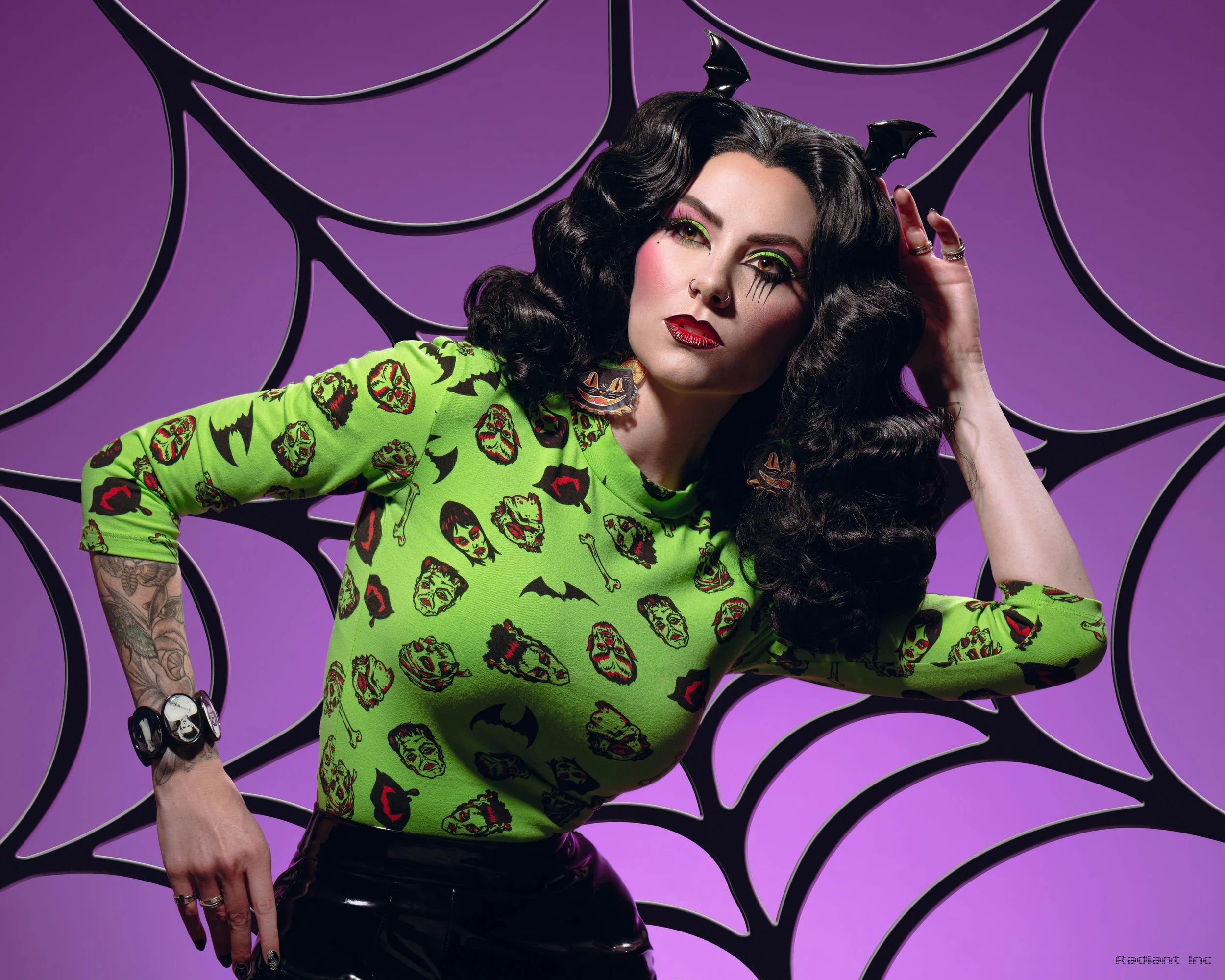 A woman with dark, curly hair, wearing a bright green shirt with cartoon monster faces, bat and bone patterns, poses in front of a purple backdrop with black web-like patterns, adding a spooky Halloween vibe.