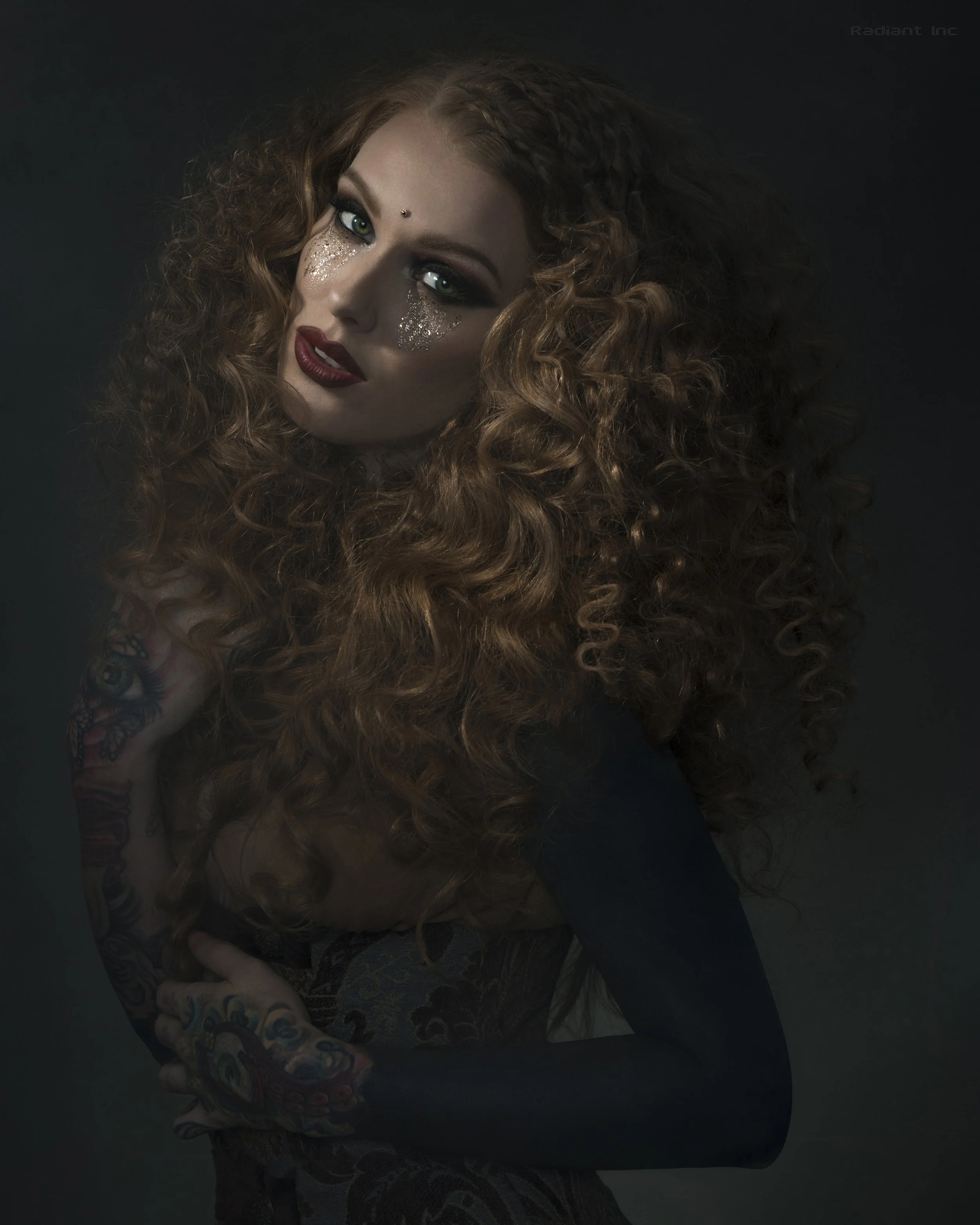 A woman with curly red hair, makeup with glitter around her eyes, and tattoos on her arms. She is looking at the camera against a dark background.