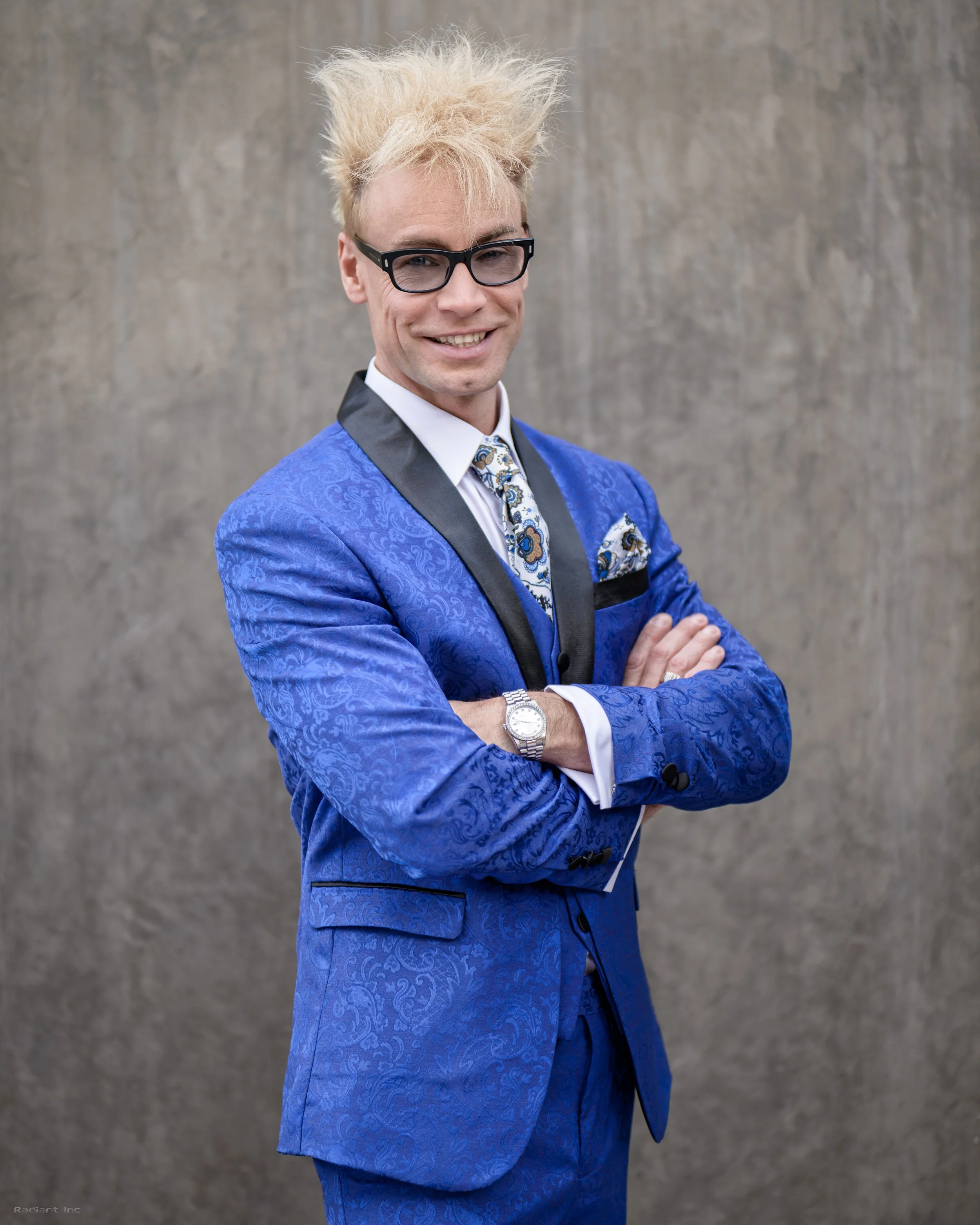 A man with blond, spiky hair, wearing black glasses and a blue patterned suit, crosses his arms and smiles against a plain background.