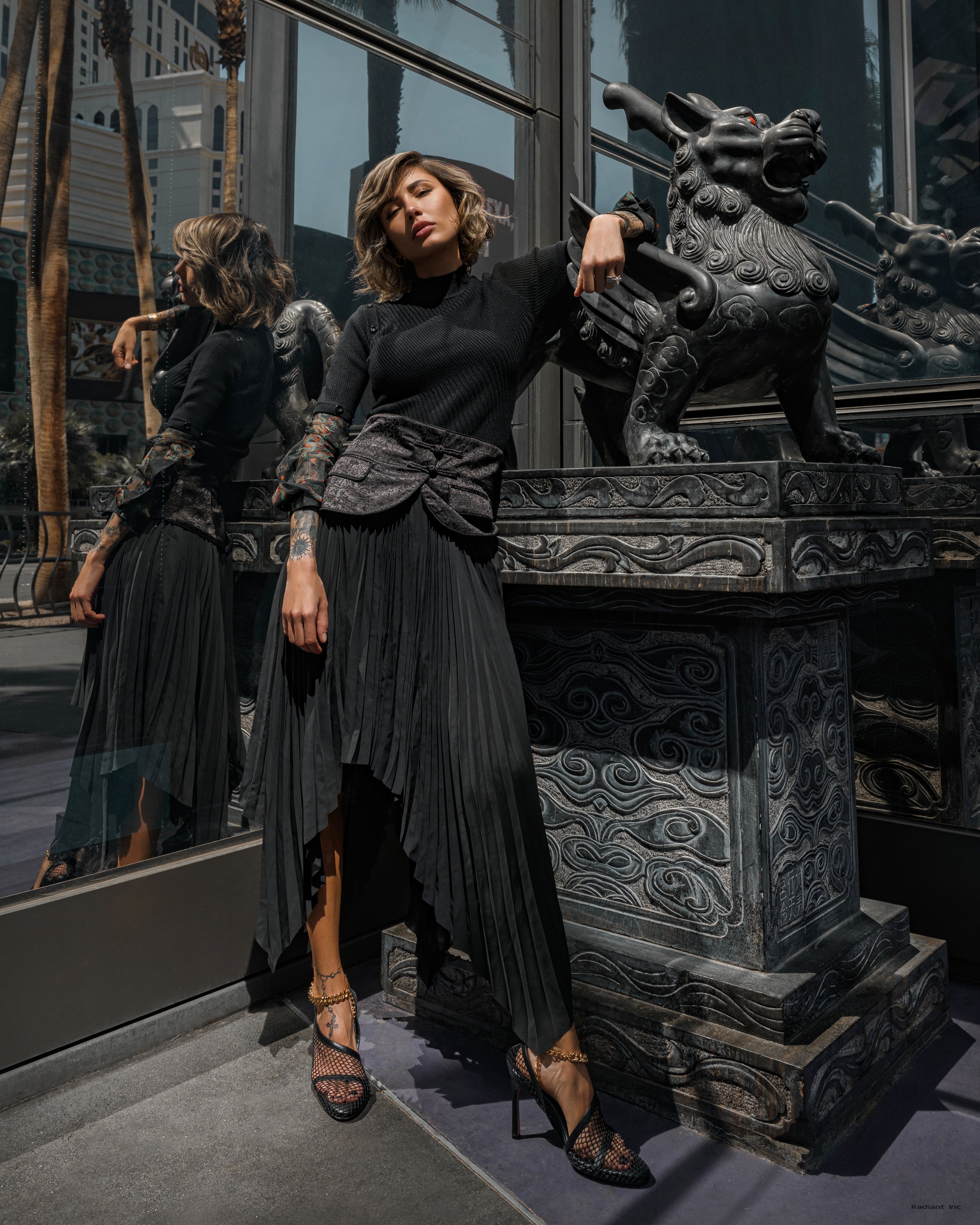 A woman with short, wavy hair in a black top and pleated black skirt standing next to a decorative Chinese guardian lion sculpture, with her arm resting on it and her reflection visible in a mirror behind her.