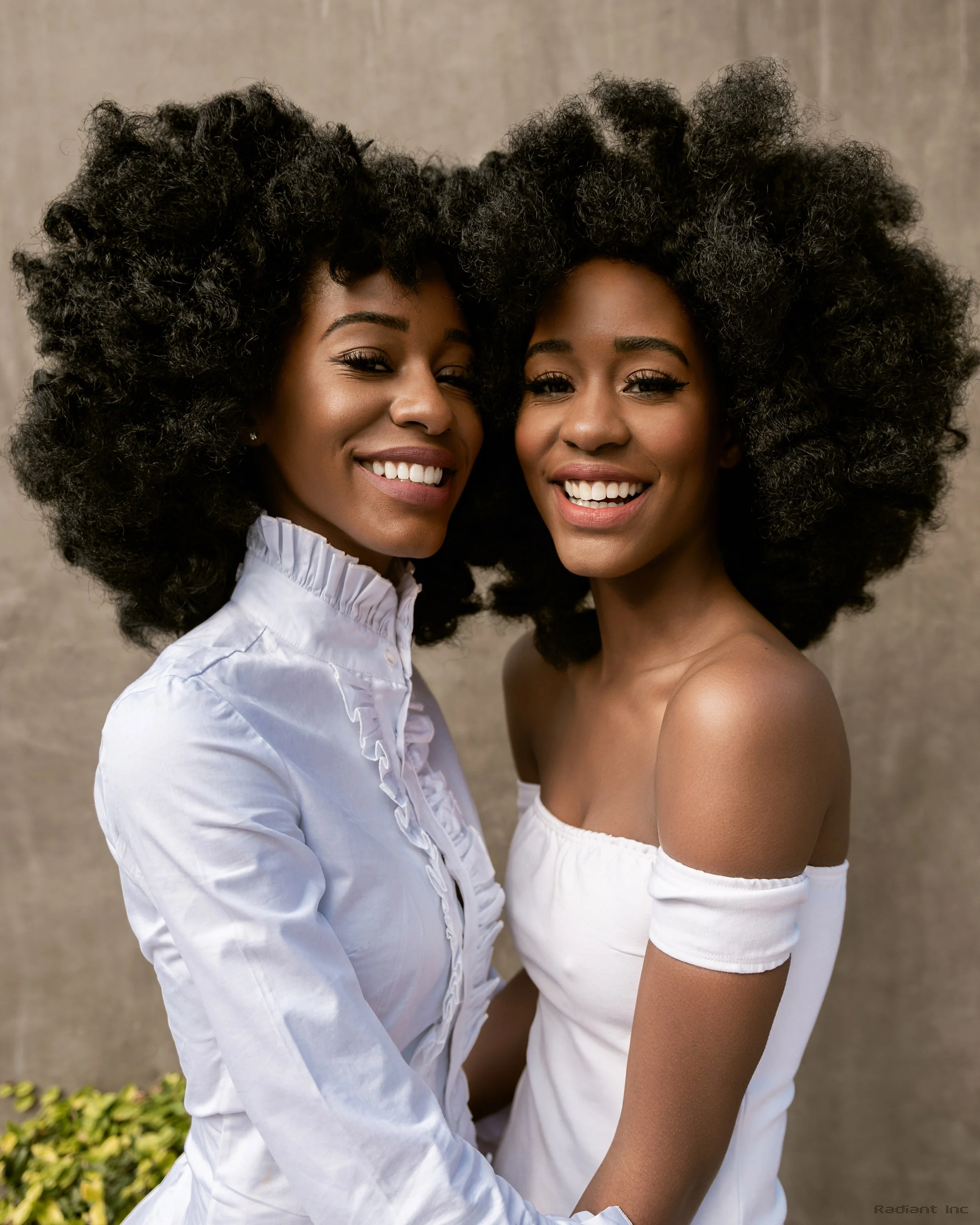 Two women with large, curly black hair smiling and embracing each other in front of a neutral background.
