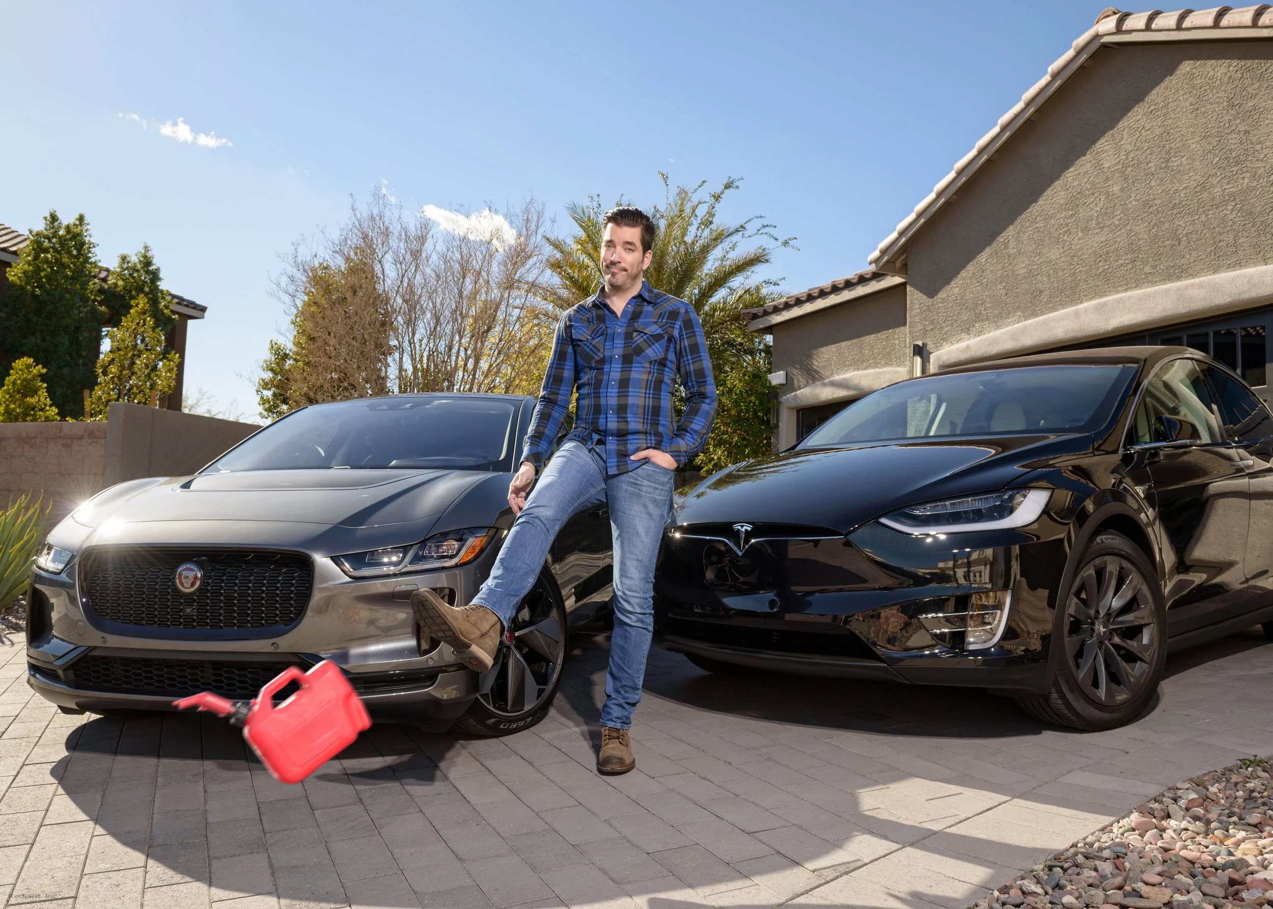A man standing with one foot on a gray Jaguar car and the other foot on a black Tesla car in a driveway outside a house, with a red gas can next to the Jaguar.