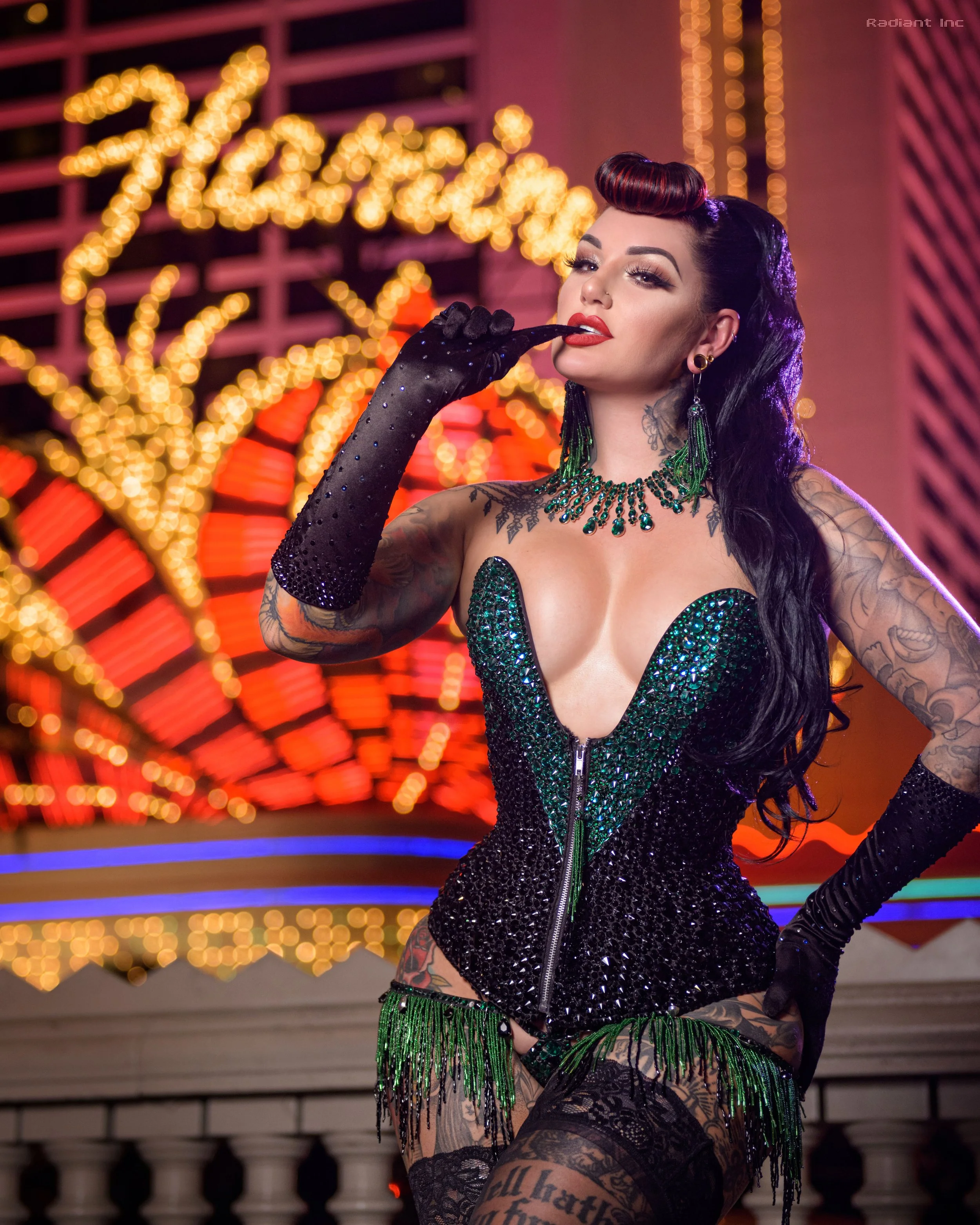 A woman with tattoos dressed in a sparkly black and green corset, black lace stockings, and long black gloves, standing in front of a bright, colorful neon-lit background.