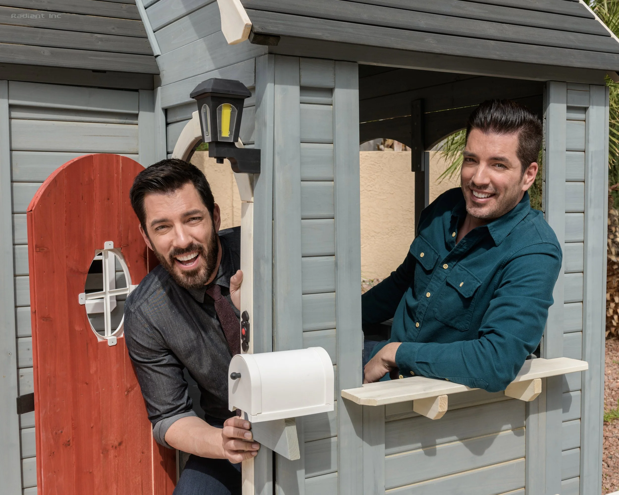 Two men with dark hair and beards smiling, one with a suit and tie, the other more casual, playing inside a wooden playhouse with a red door and gray walls, outdoors on a sunny day.
