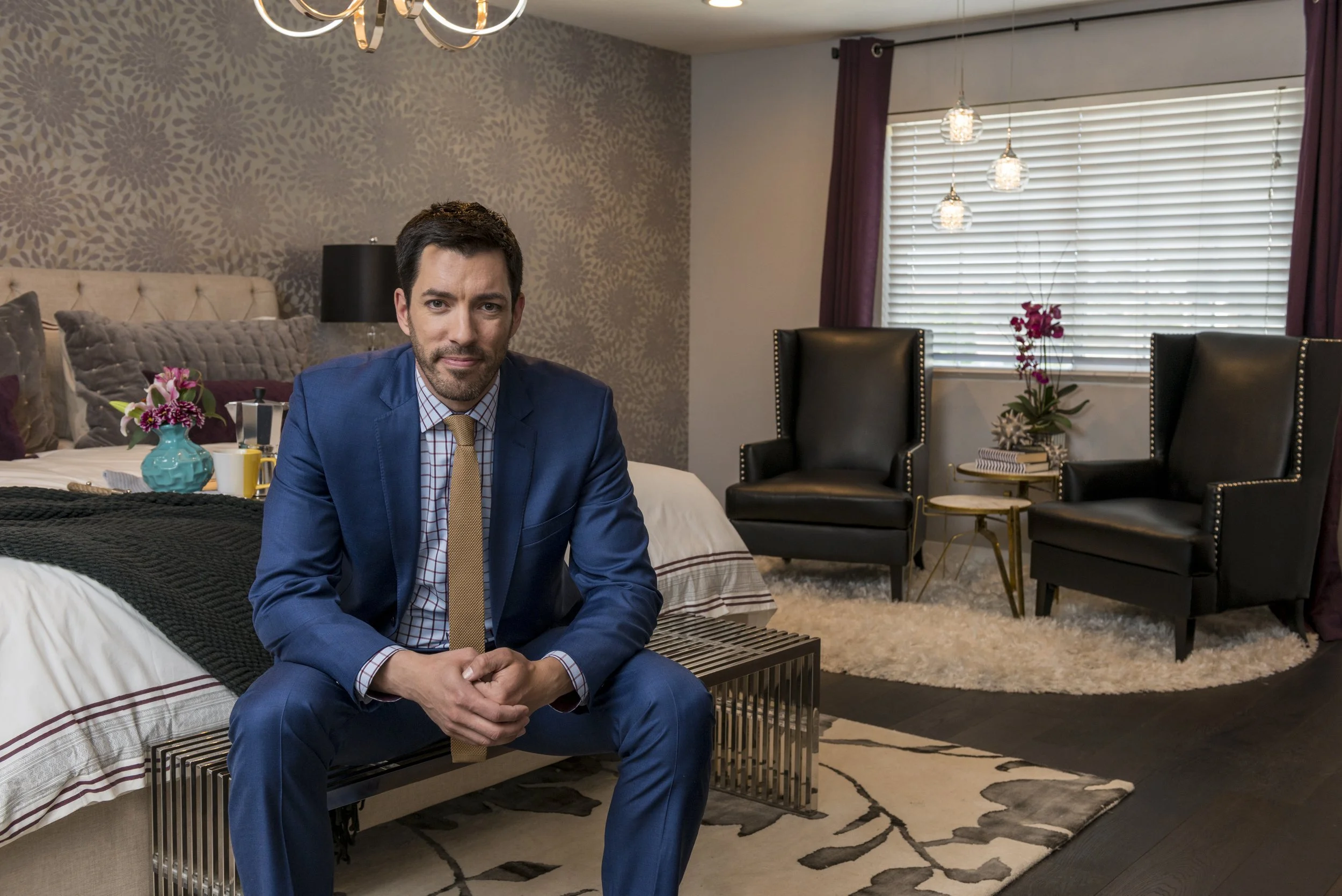 A man in a blue suit sitting on a bench at the edge of a bed in a bedroom with floral wallpaper, a large bed with pillows and a blanket, and black armchairs by a window with purple curtains and hanging lights.