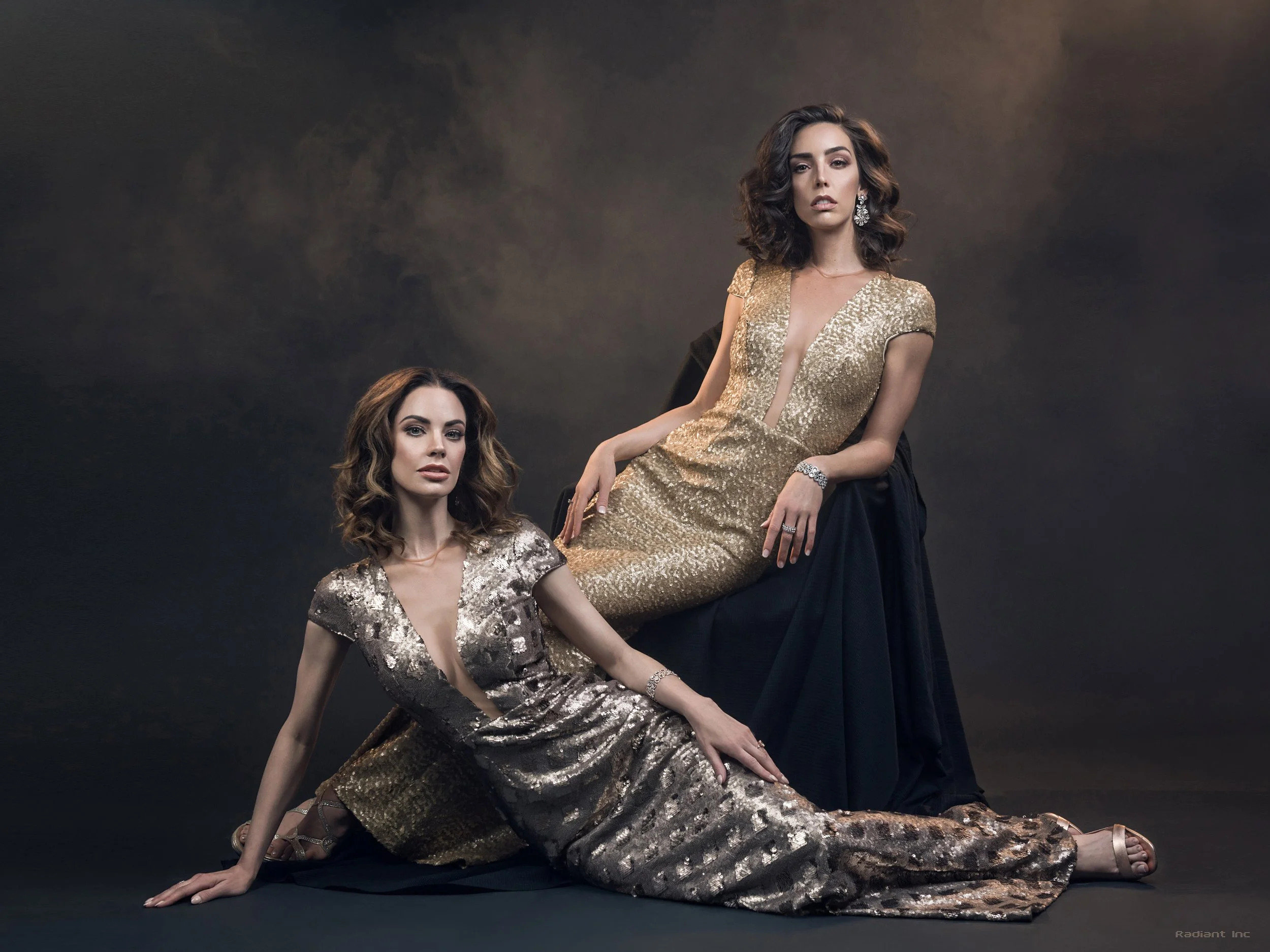 Two women in glamorous gold and silver sequin dresses pose against a dark background, one sitting on the floor with her hand on the ground and the other sitting on a high black stool with a draped black cloth behind her.