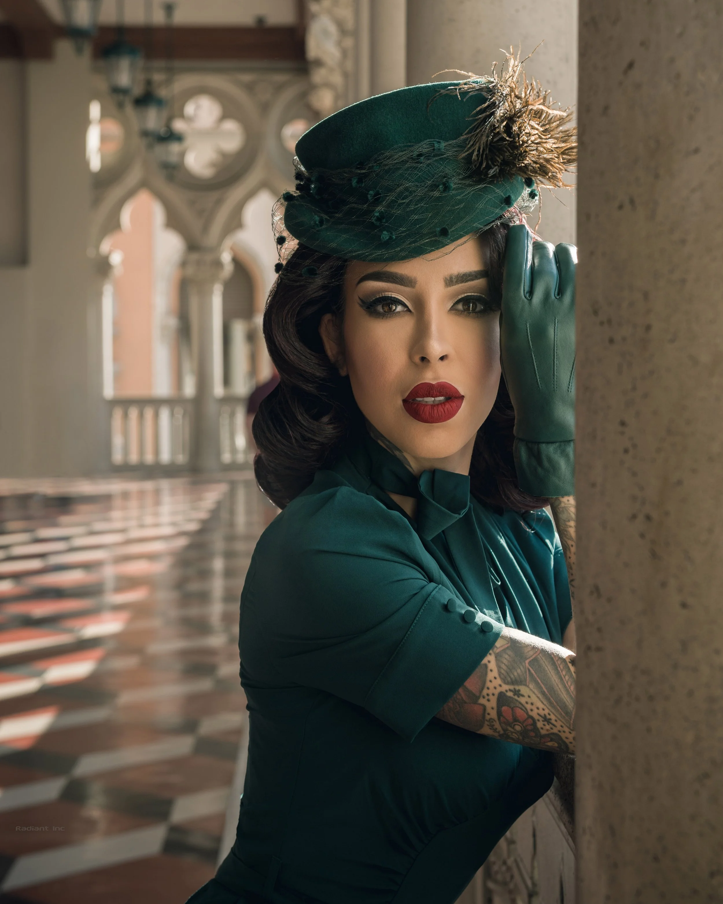 A woman with dark hair, red lipstick, and tattooed arms, dressed in vintage style clothing and a green hat with feathers, standing by a stone column in an elegant interior.