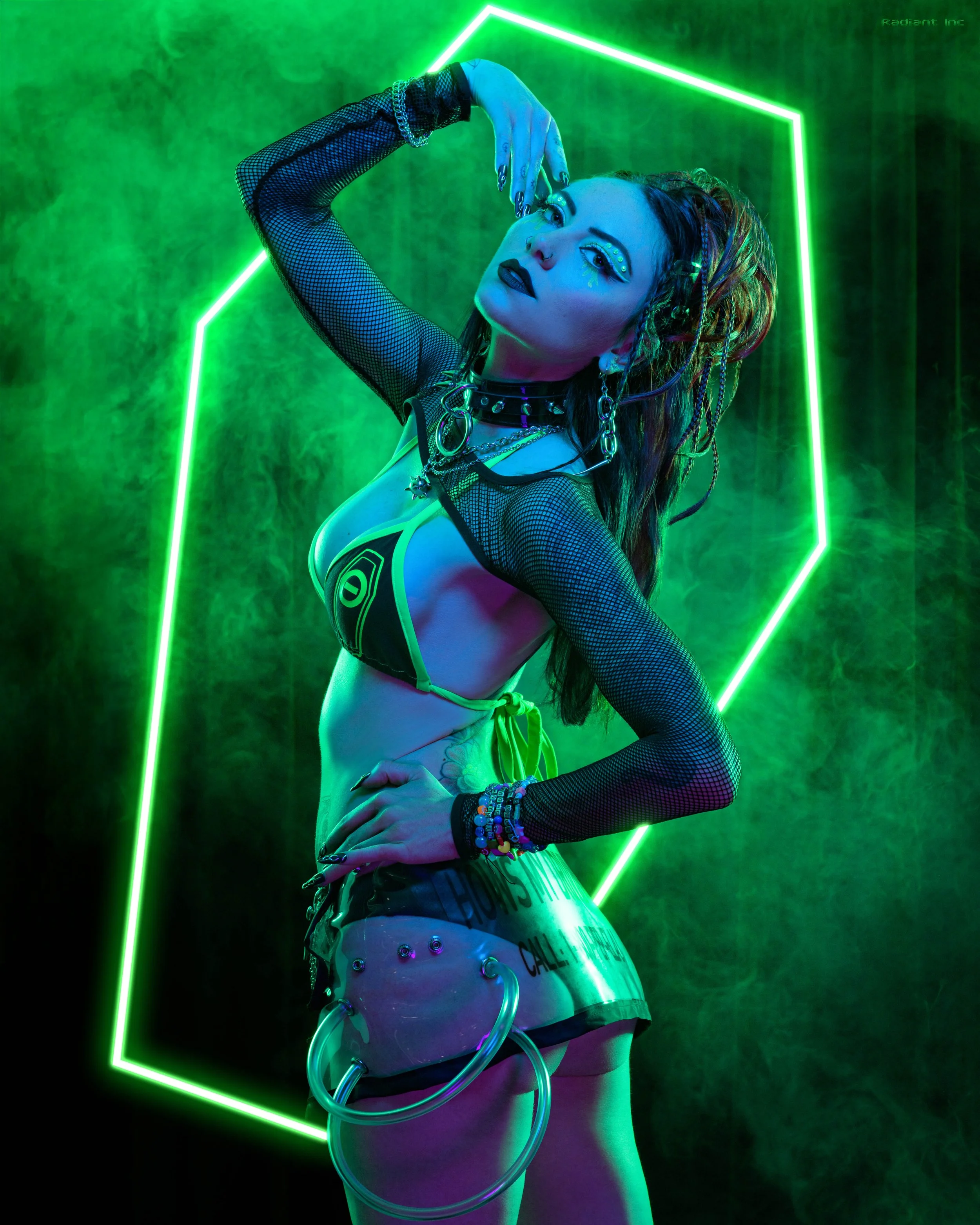 A woman in cyberpunk-styled attire with fishnet arm sleeves, chains, and colorful accessories posing confidently in front of green neon geometric shapes with smoke effects.