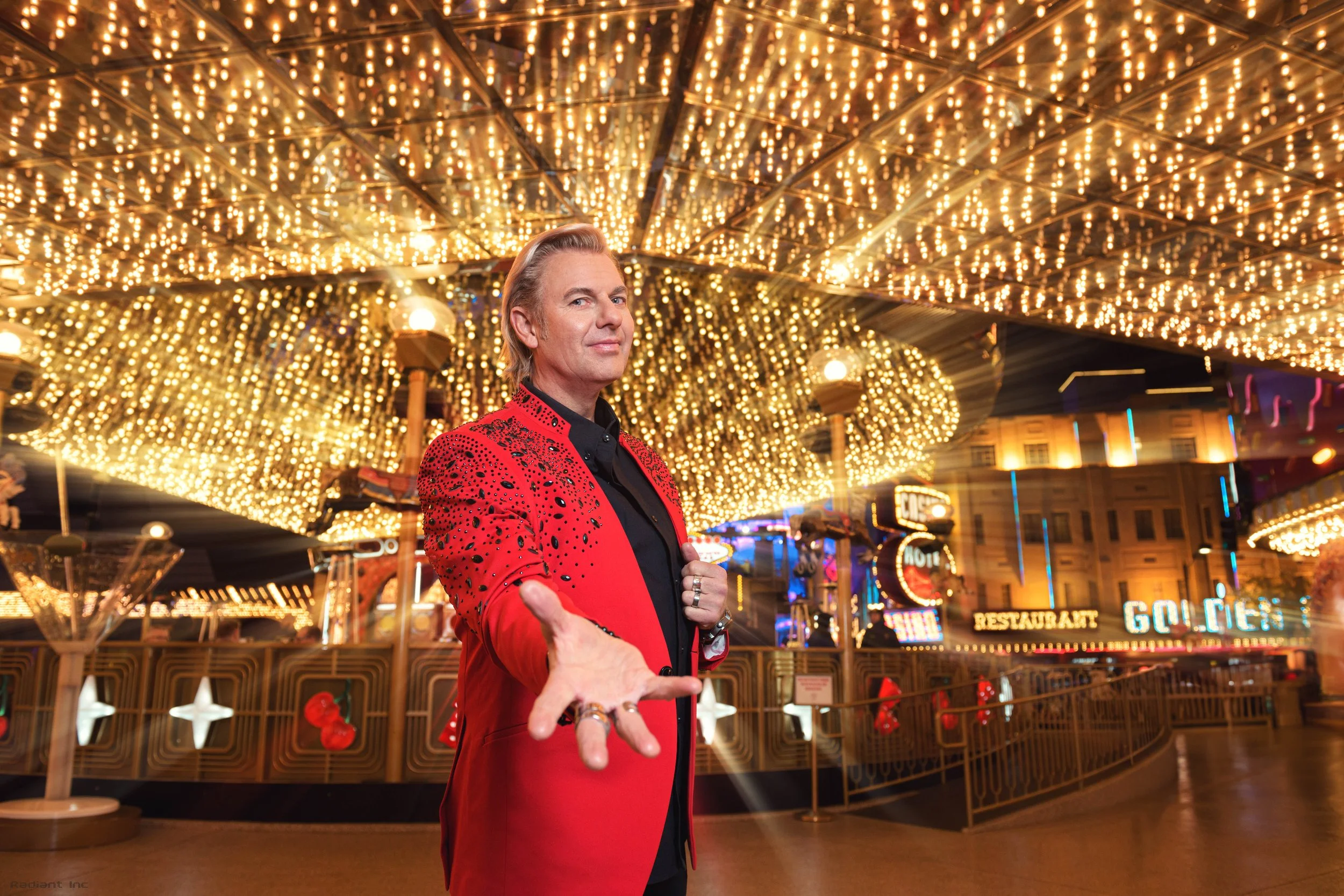 A man in a red jacket with black spots extending his hand towards the camera, standing under a canopy of bright, golden string lights at a festive amusement park or carnival.