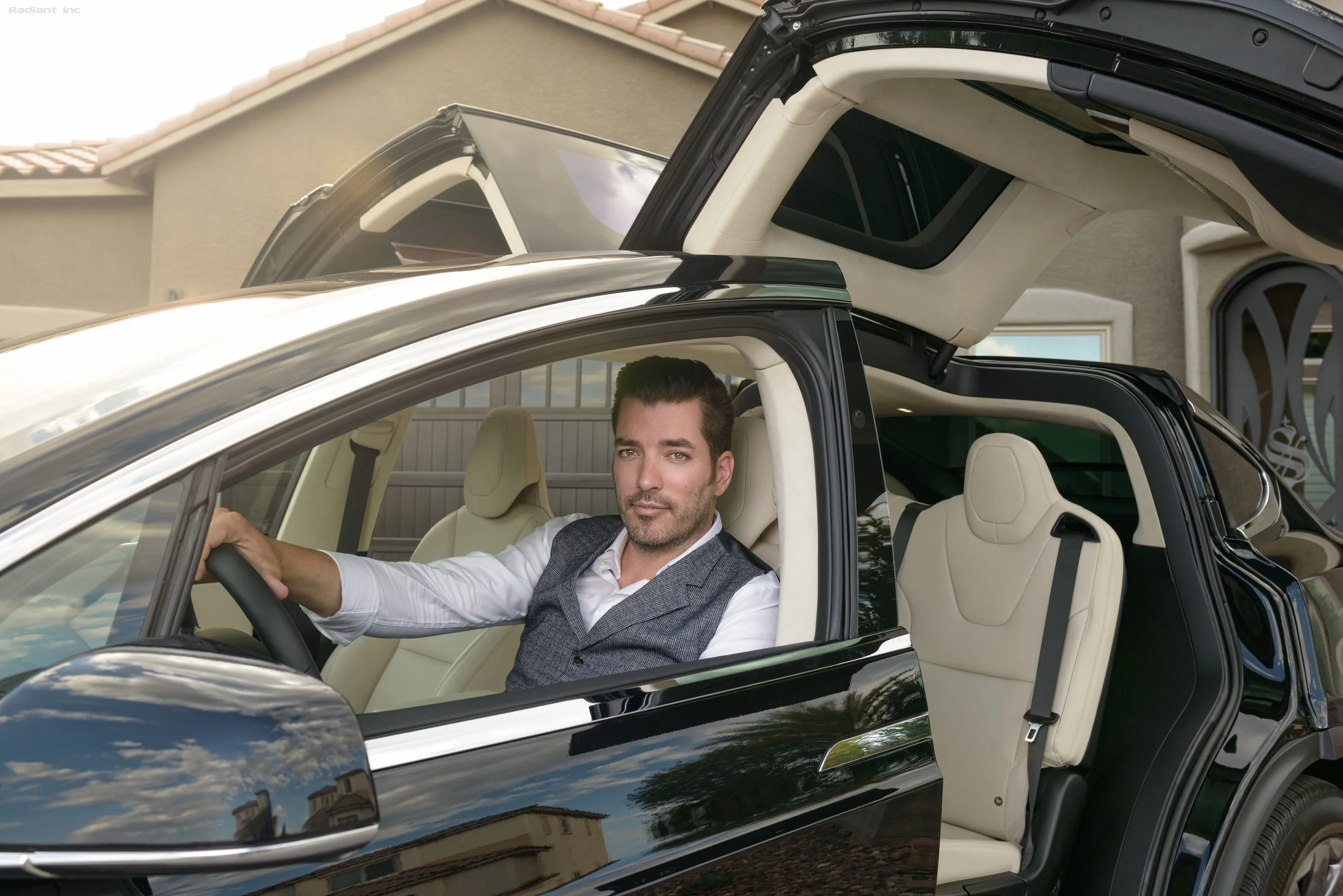 A man sitting in the driver's seat of a black luxury SUV with its gullwing doors open, in front of a house.