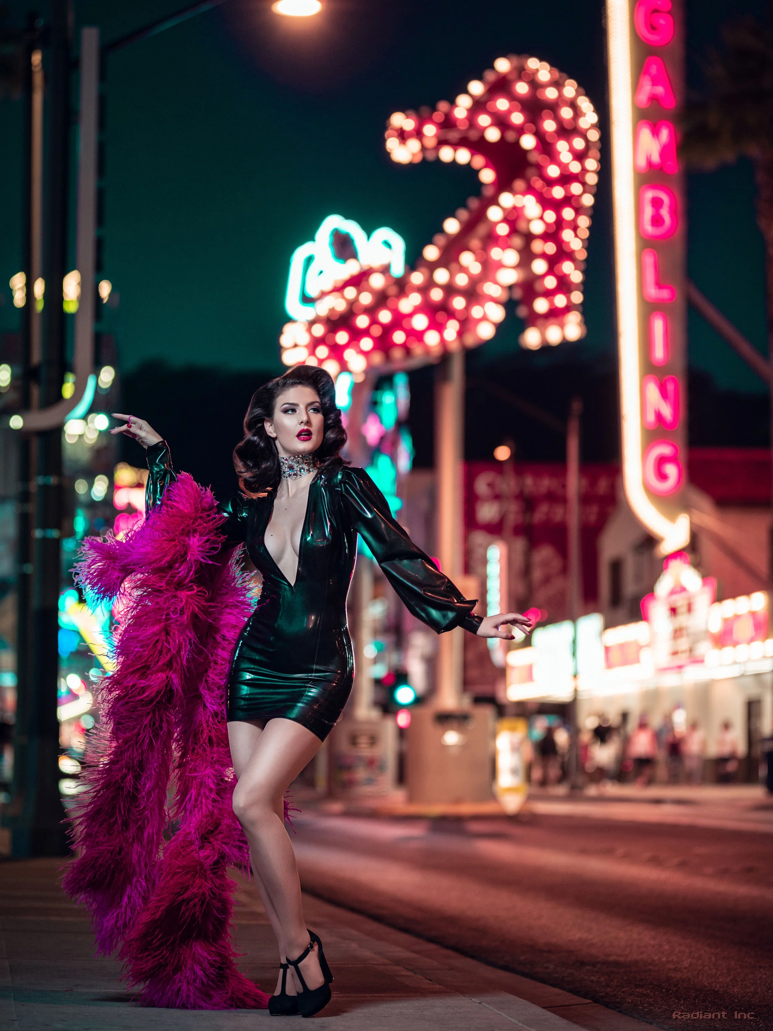 A woman in a black shiny dress with a pink feather boa walking on a city street at night with neon signs, including a large game sign in the background.