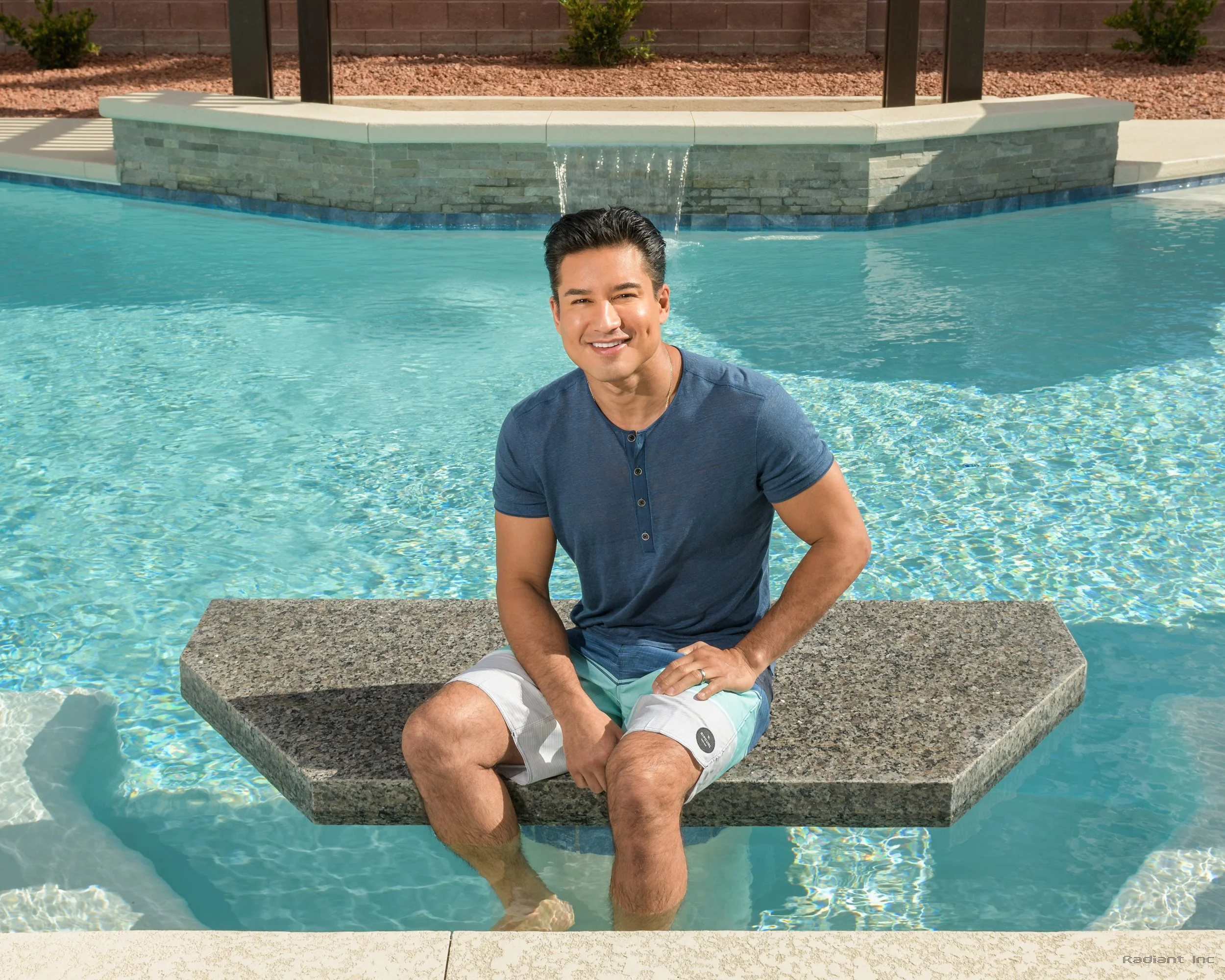 A man sitting on a stone bench in a swimming pool, smiling at the camera with water up to his ankles, wearing a blue t-shirt and light-colored shorts, with a fountain and landscaped area in the background.