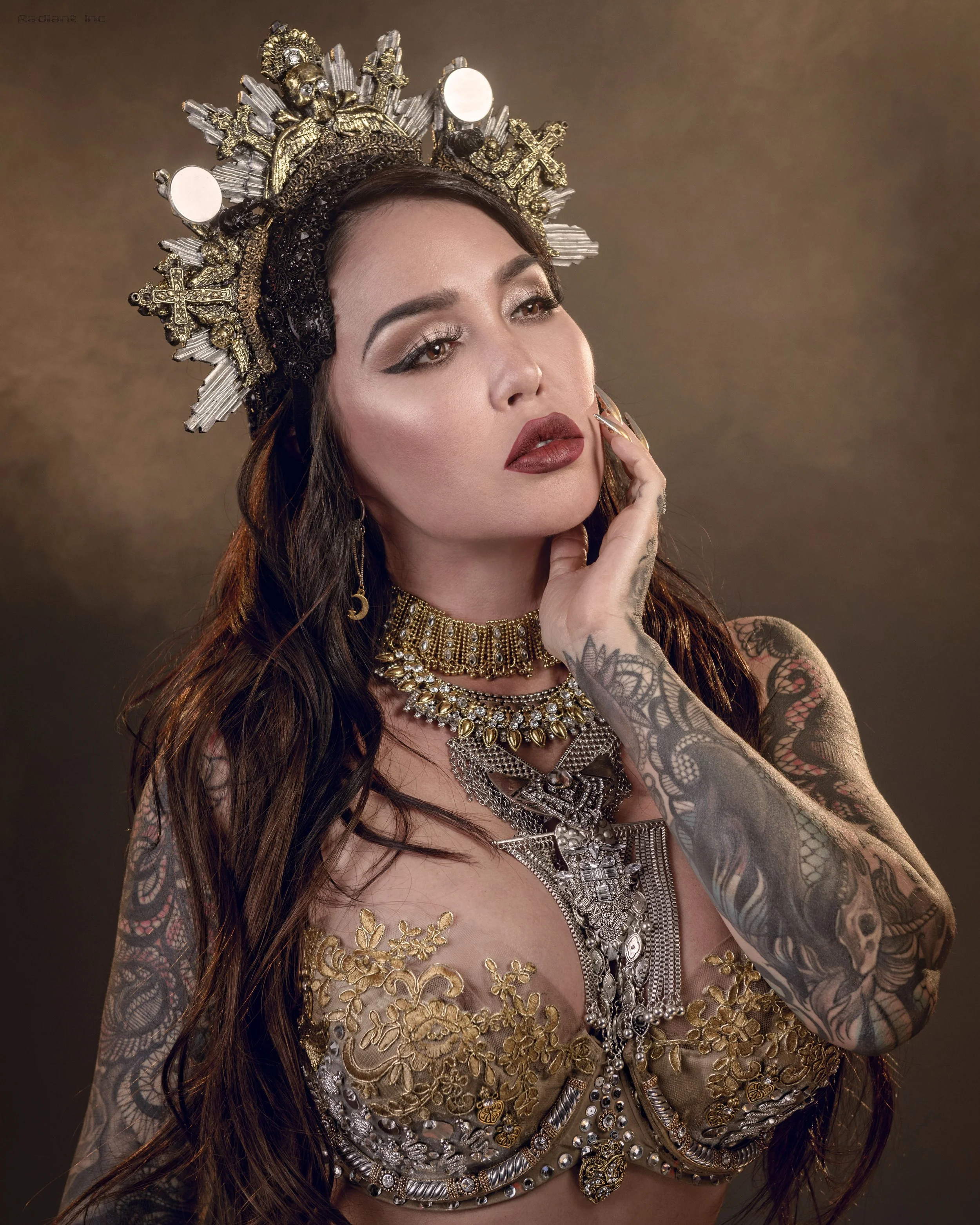 A woman with long brown hair and tattoos wearing gold and silver jewelry, including a crown-like headpiece, in front of a brown background.