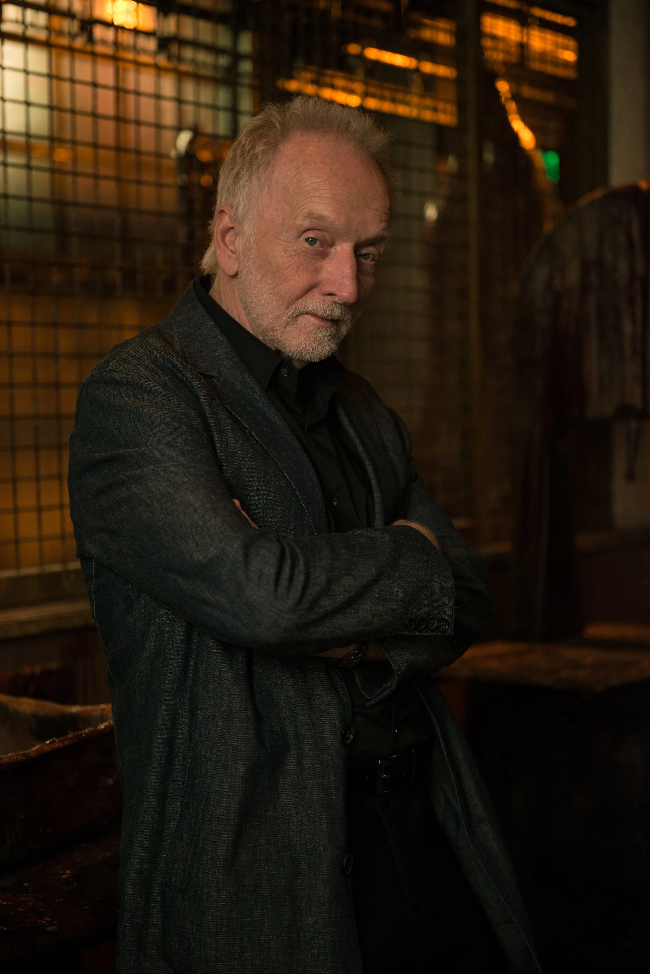 An older man with a beard and mustache, wearing a dark blazer and shirt, standing with arms crossed in a dimly lit room with a grid-like wall and warm lighting.