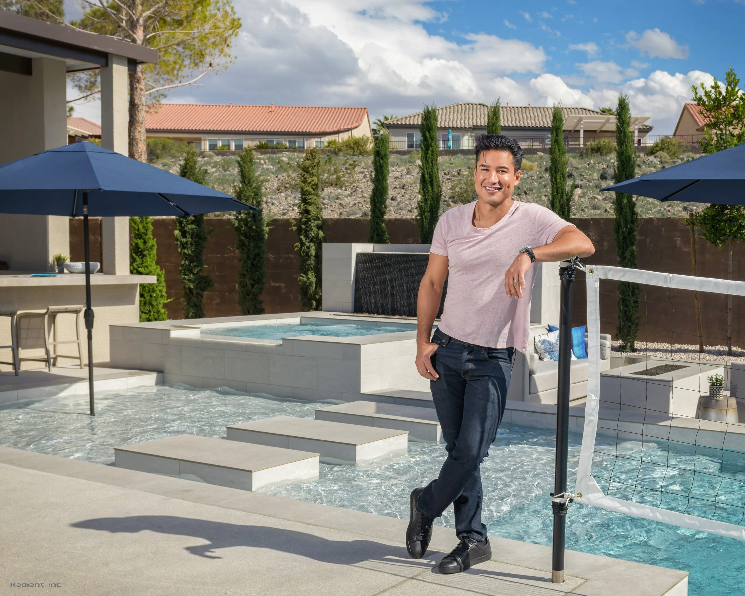 A man standing by a poolside in a backyard, smiling at the camera, with a volleyball net and umbrellas in the background.