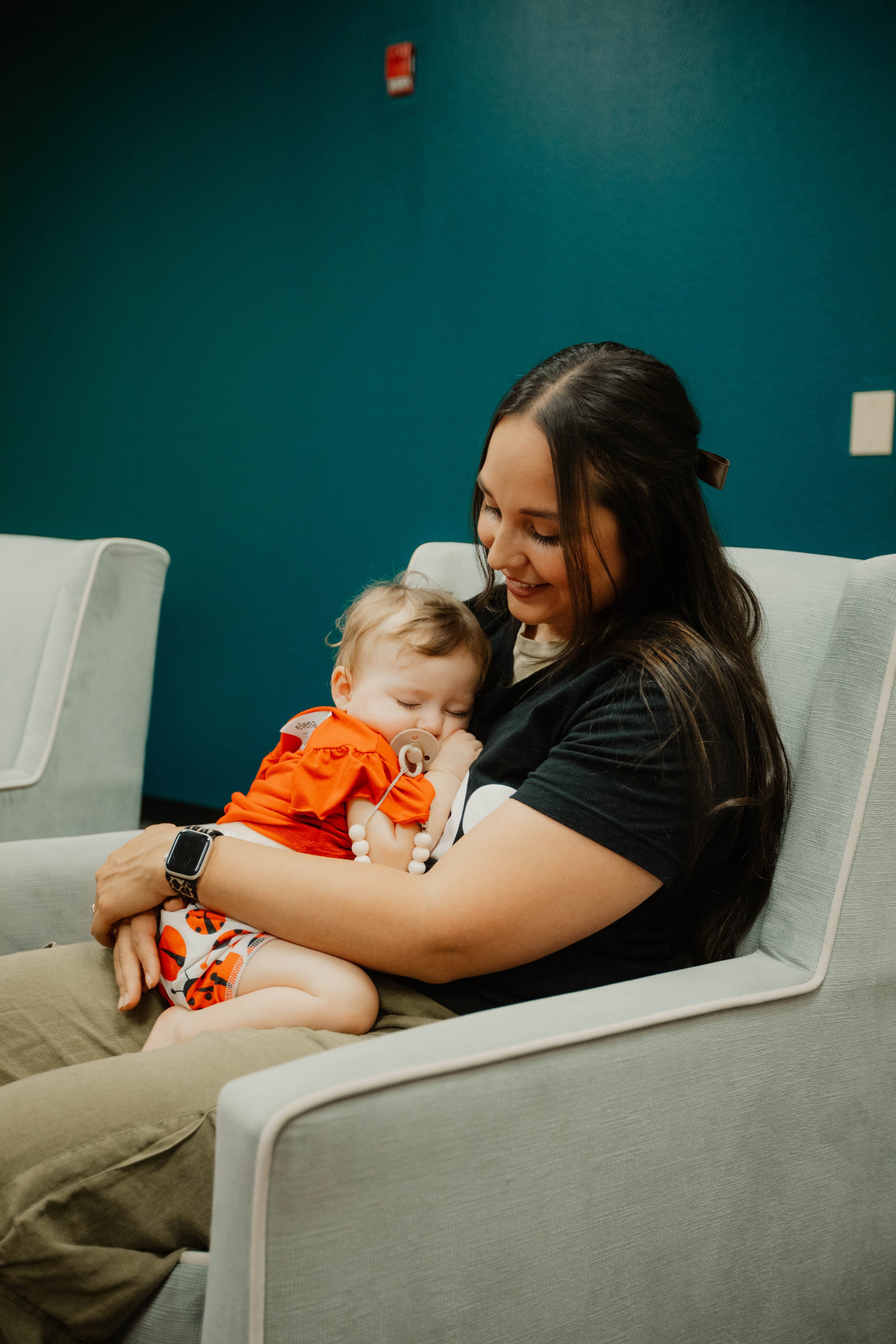 Woman holding a sleeping baby with a pacifier, sitting on a light gray chair in a room with blue walls.