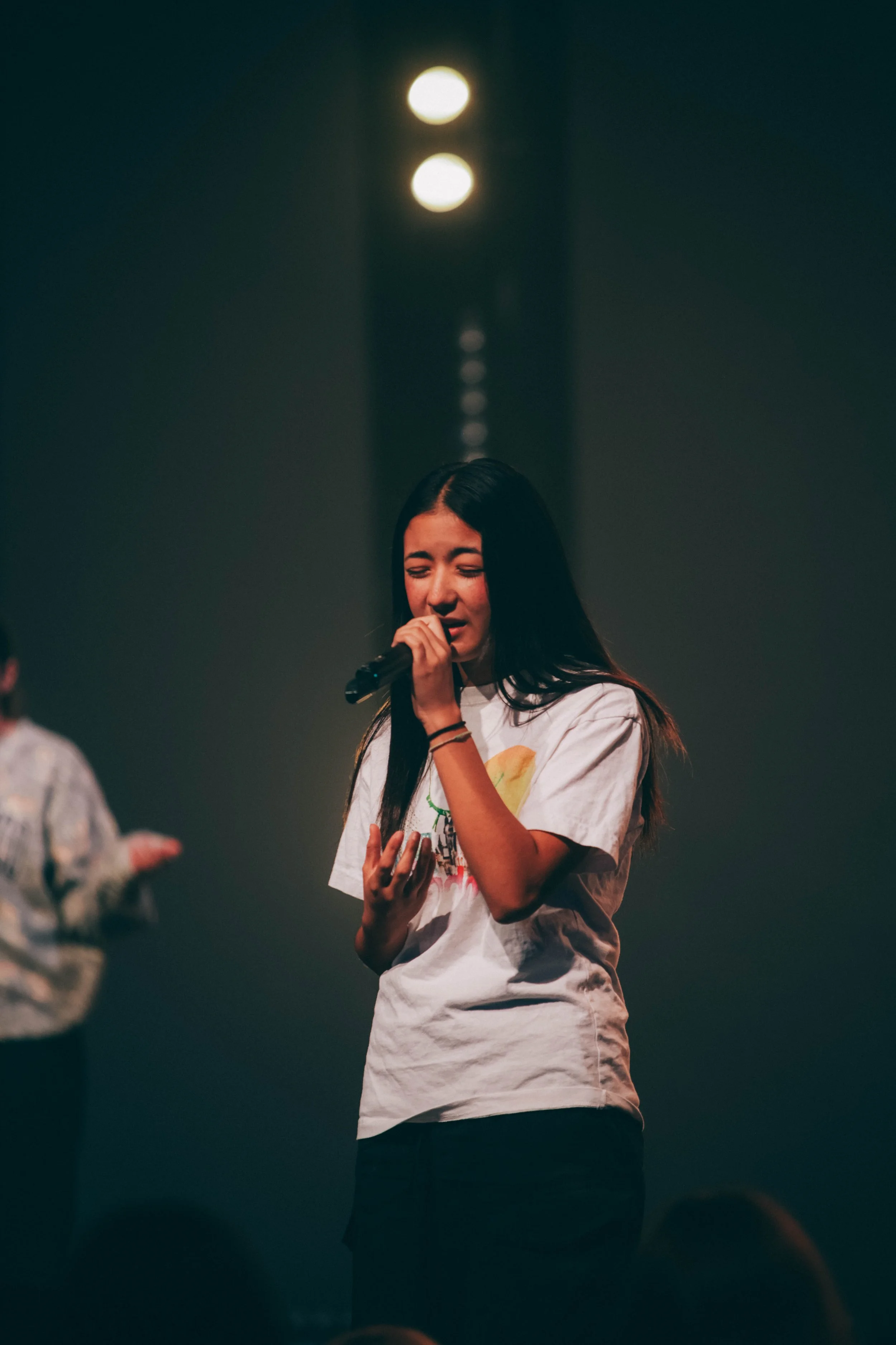 A young woman with long dark hair singing into a microphone on stage with stage lights behind her.