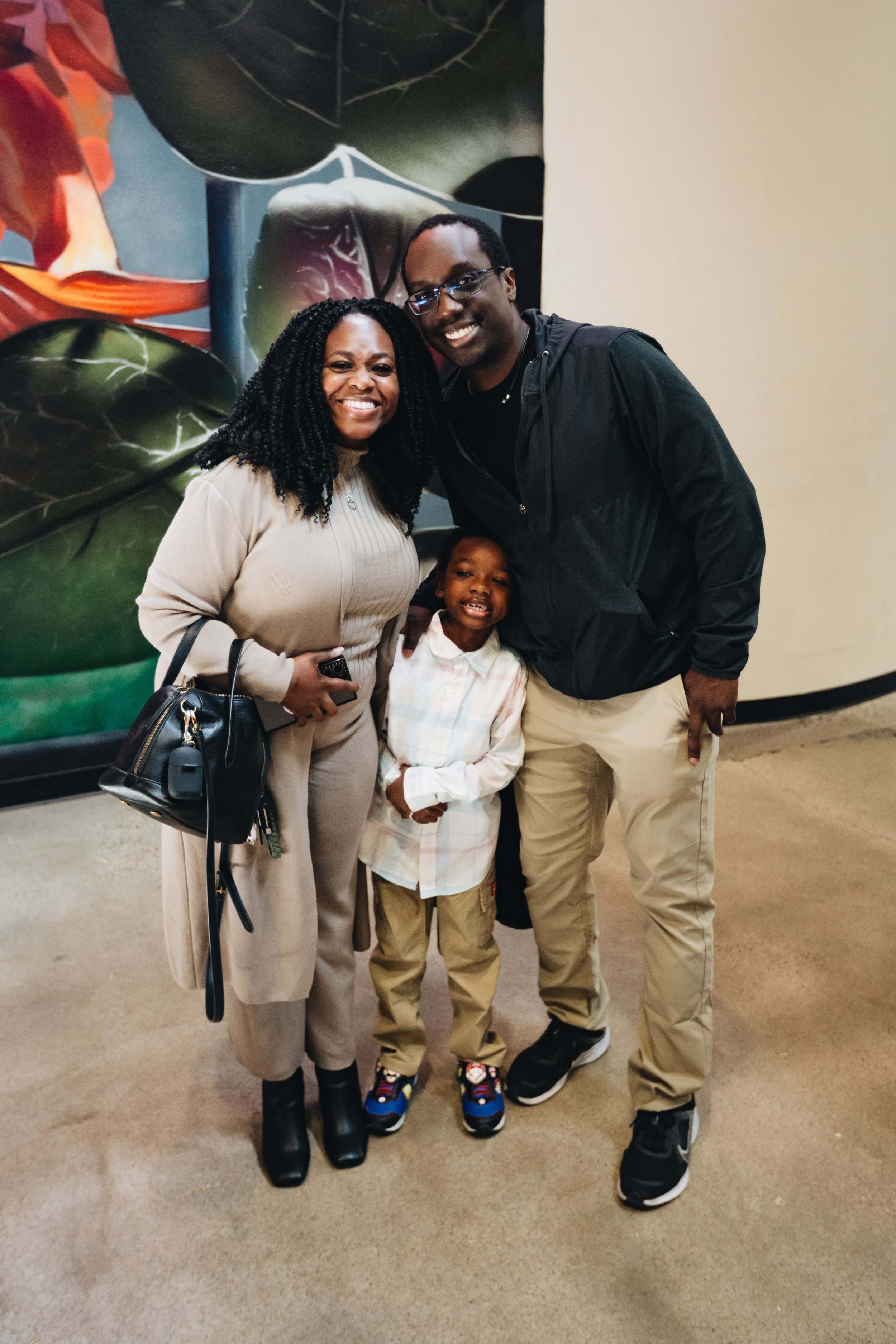 A family of three, a woman, a man, and a young boy, smiling and posing together indoors, with a colorful abstract mural in the background.