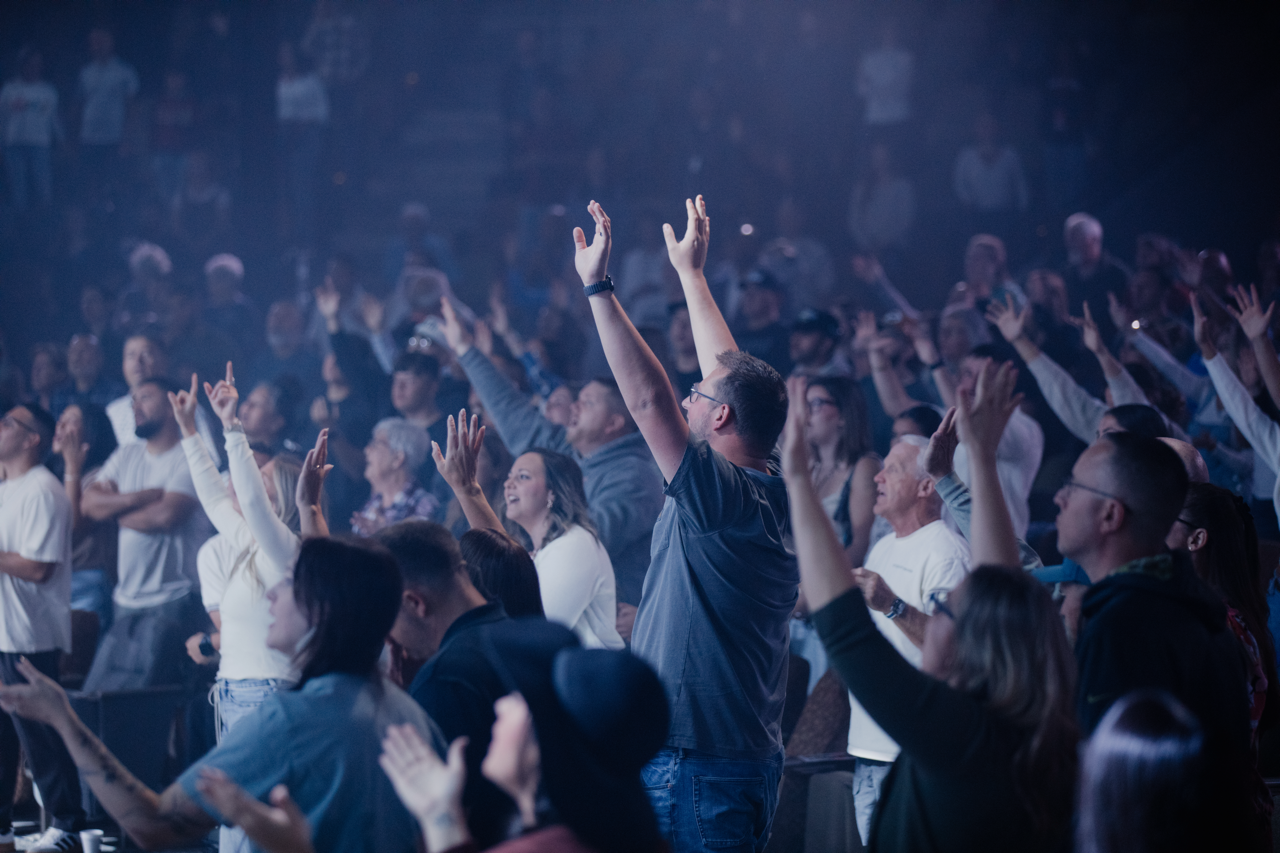 Audience at a concert or event, with some people raising their hands and others looking towards the stage, in a dimly lit venue.