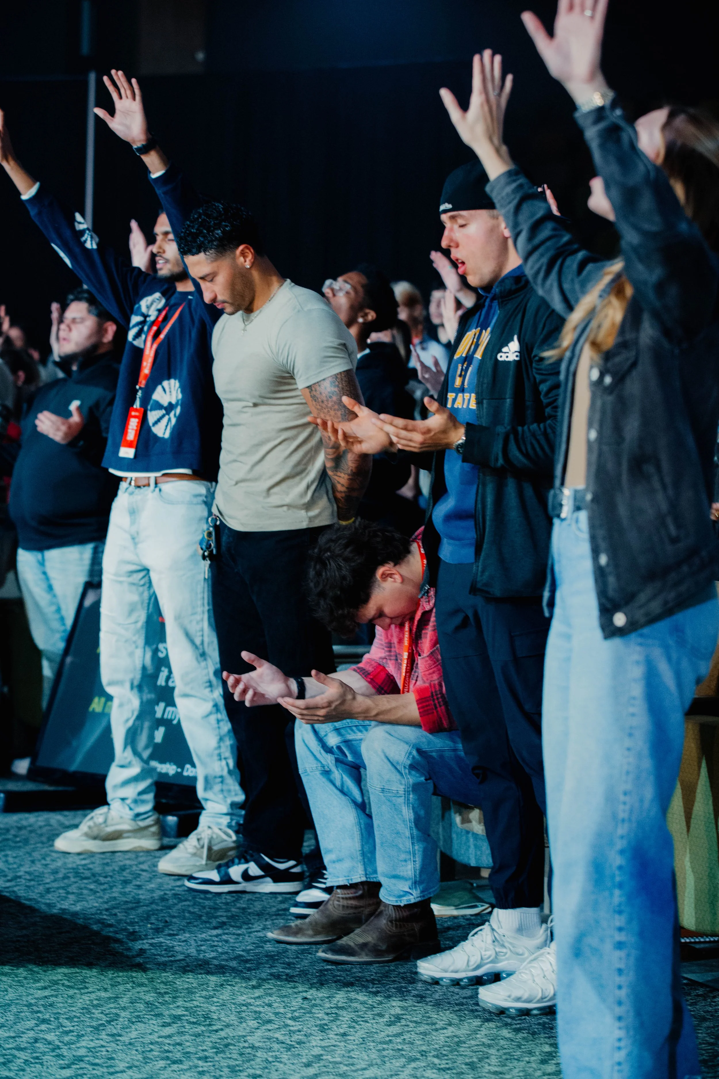 People standing and kneeling with eyes closed and hands raised during prayer or worship at a conference.