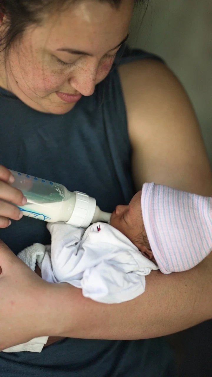 A woman holding a newborn baby and feeding it with a bottle. The baby is wearing a pink and blue striped hat and a white onesie. The woman has freckles and is wearing a dark shirt.