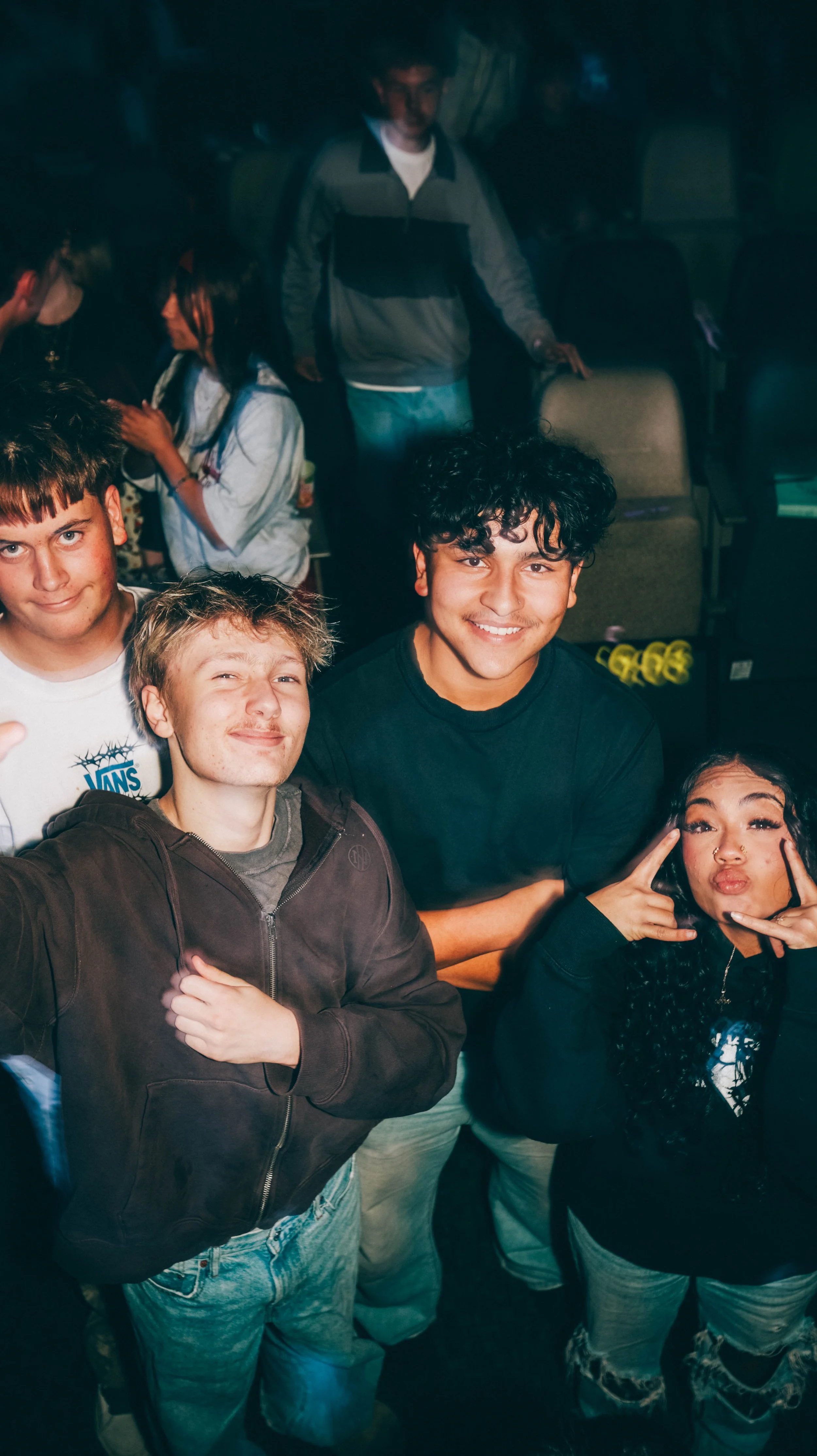 Group of young people posing for a photo, smiling and making gestures in a dark indoor setting, possibly at a social gathering or event.