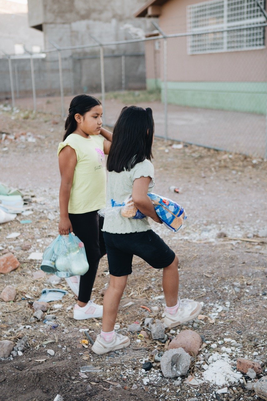 Two young girls walking on dirt ground with debris and rocks, carrying grocery bags in a rundown outdoor area near a fenced building.