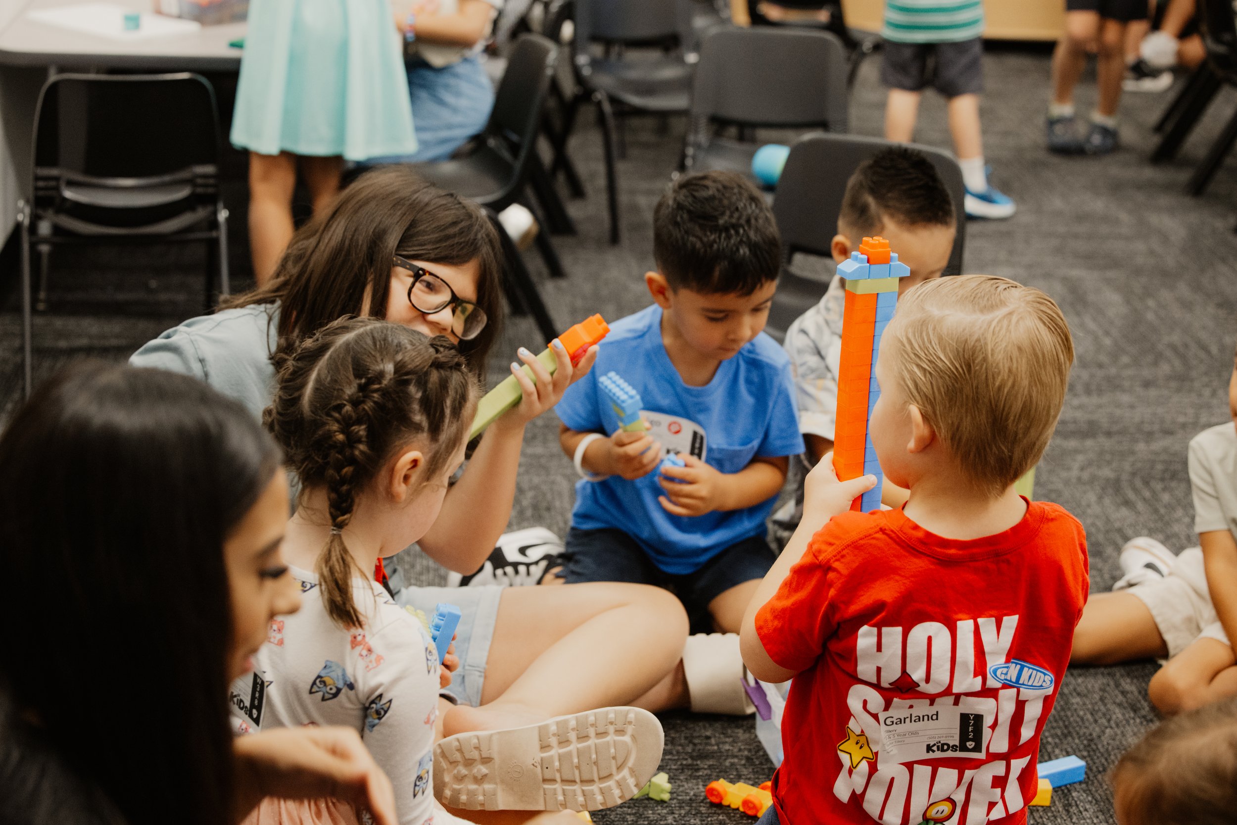 Children sitting on the floor engaging in a building activity with colorful toy blocks, with a woman supervising and helping them.