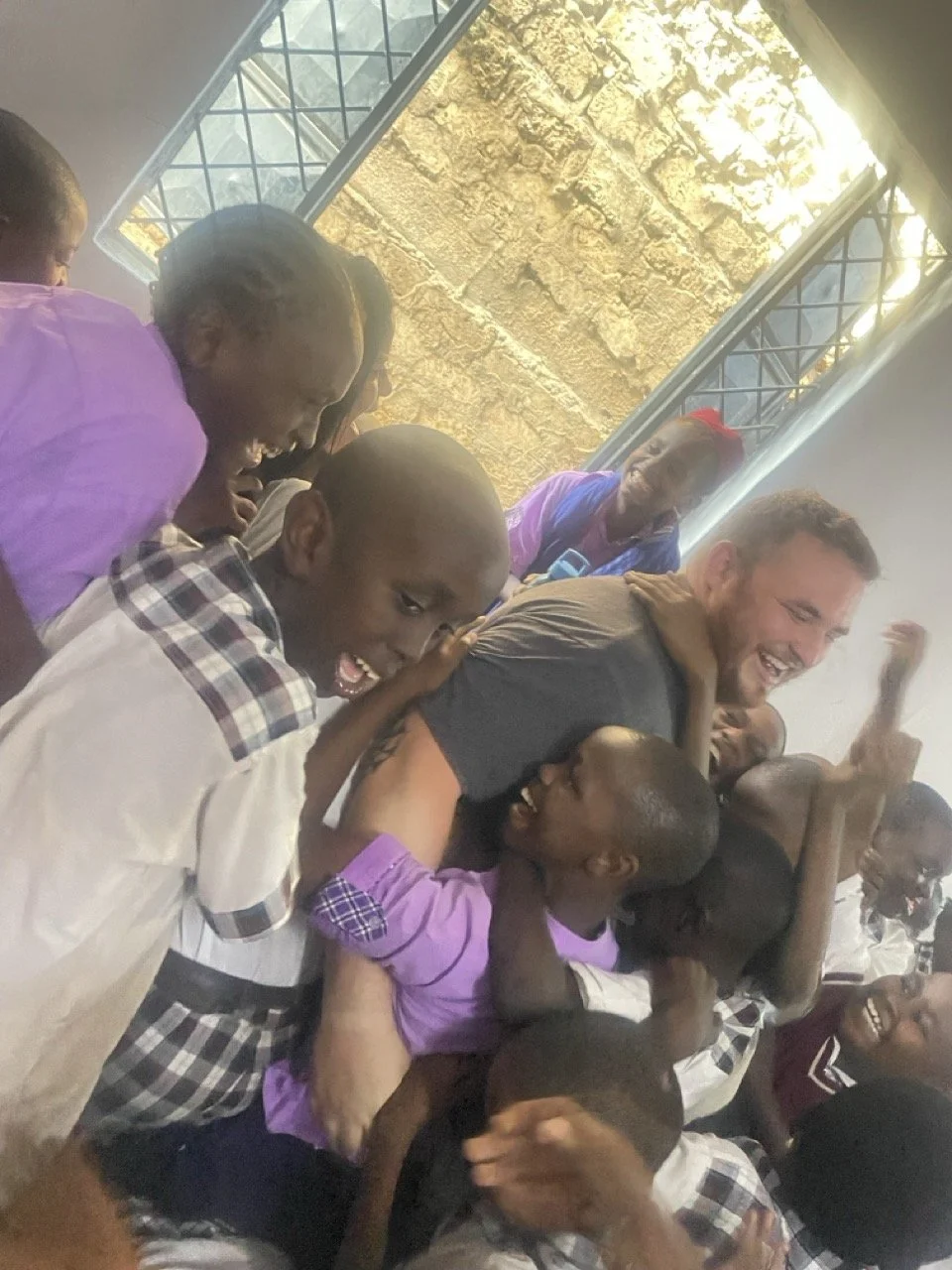 A group of children and a man are playing and laughing together indoors, with sunlight coming through a window.