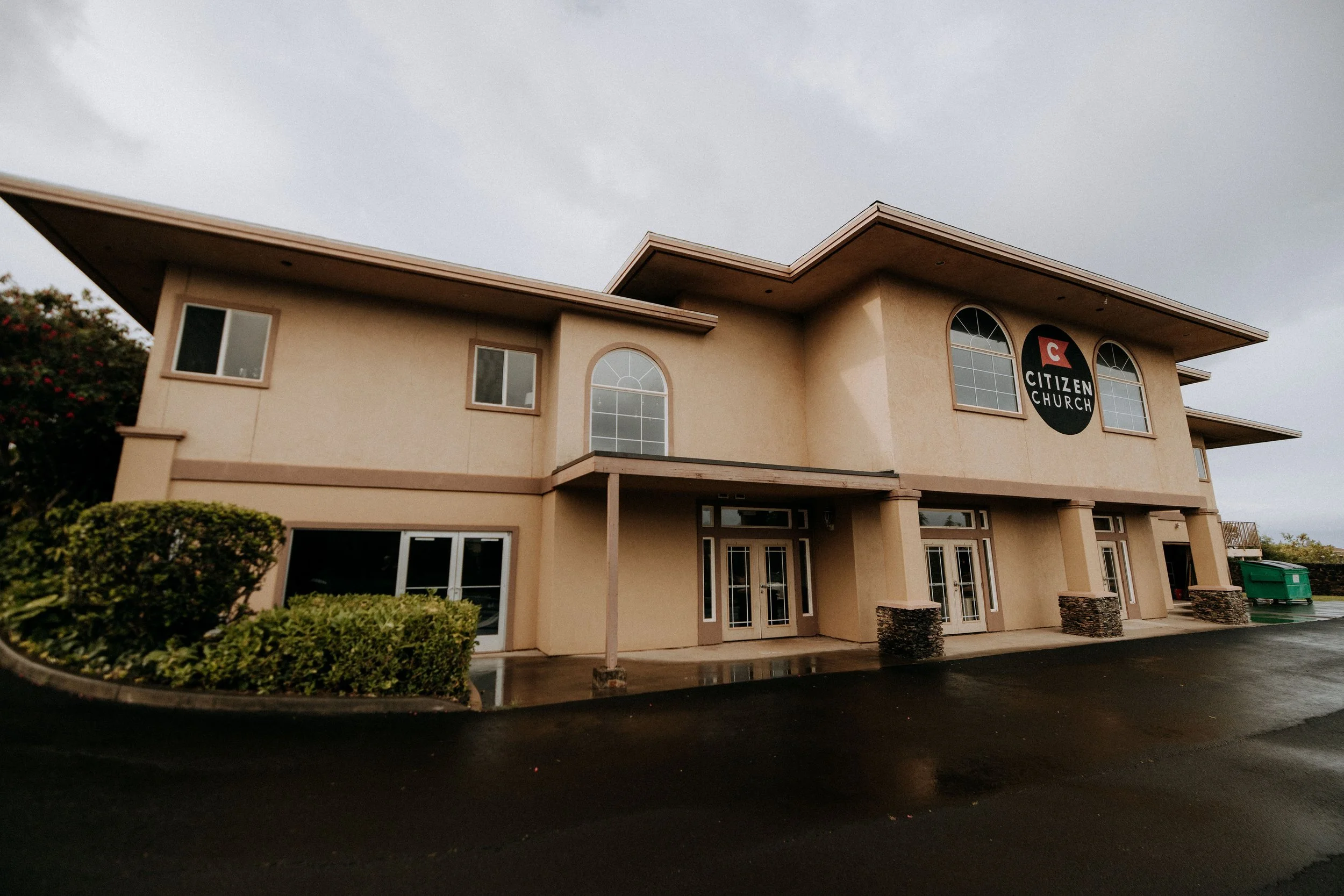 Exterior of a two-story church building with a sign that reads "Citizen Church" on the front and surrounded by shrubbery and a paved parking area.
