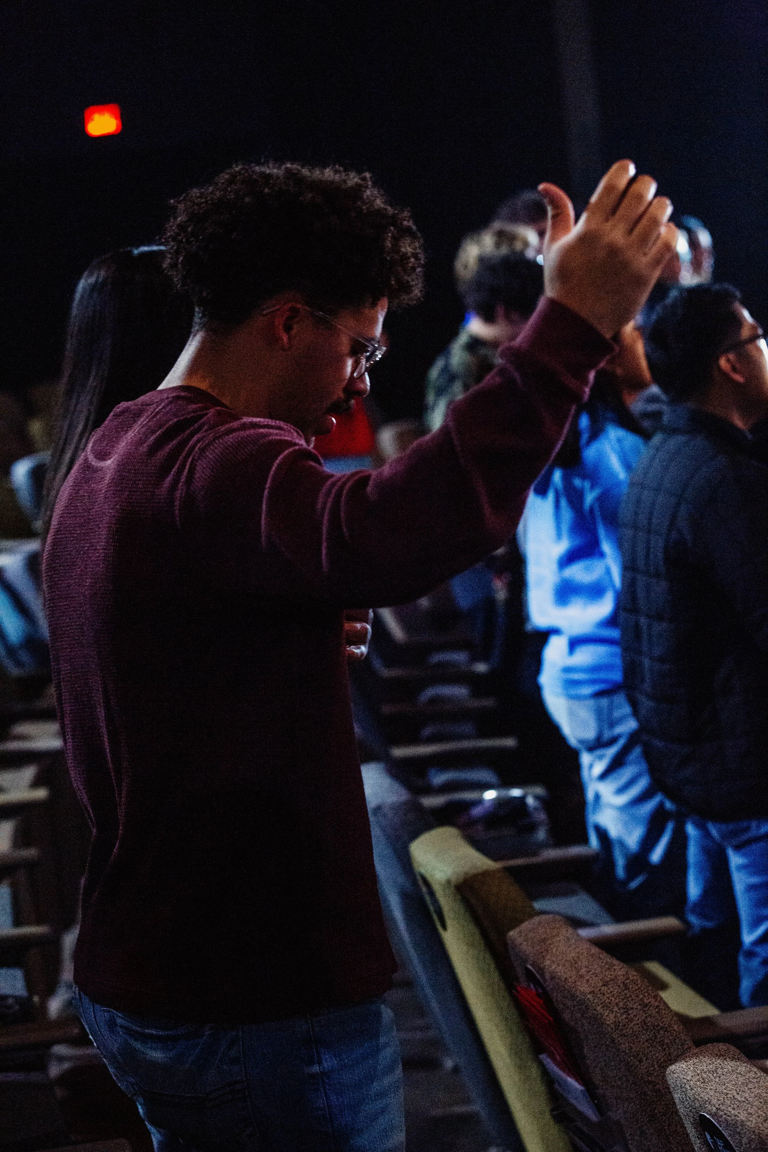 A person with curly hair and glasses praying with eyes closed in a dark auditorium.