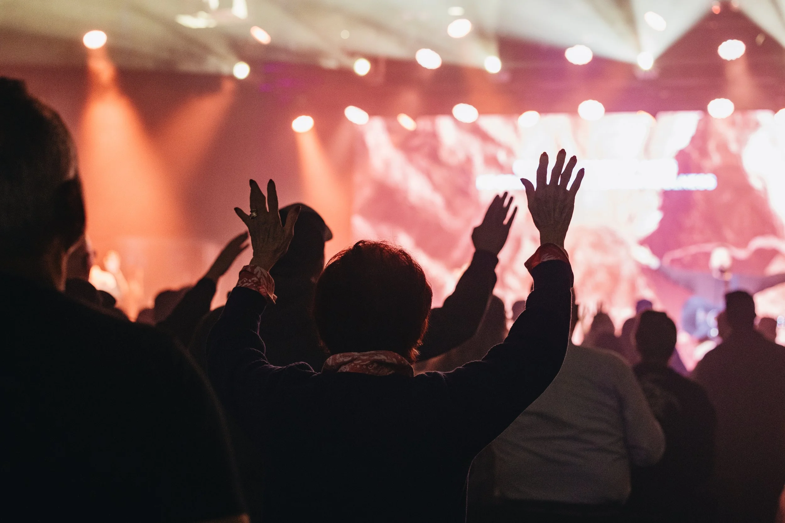 Audience raising hands during a concert or event with stage lights and colorful background.
