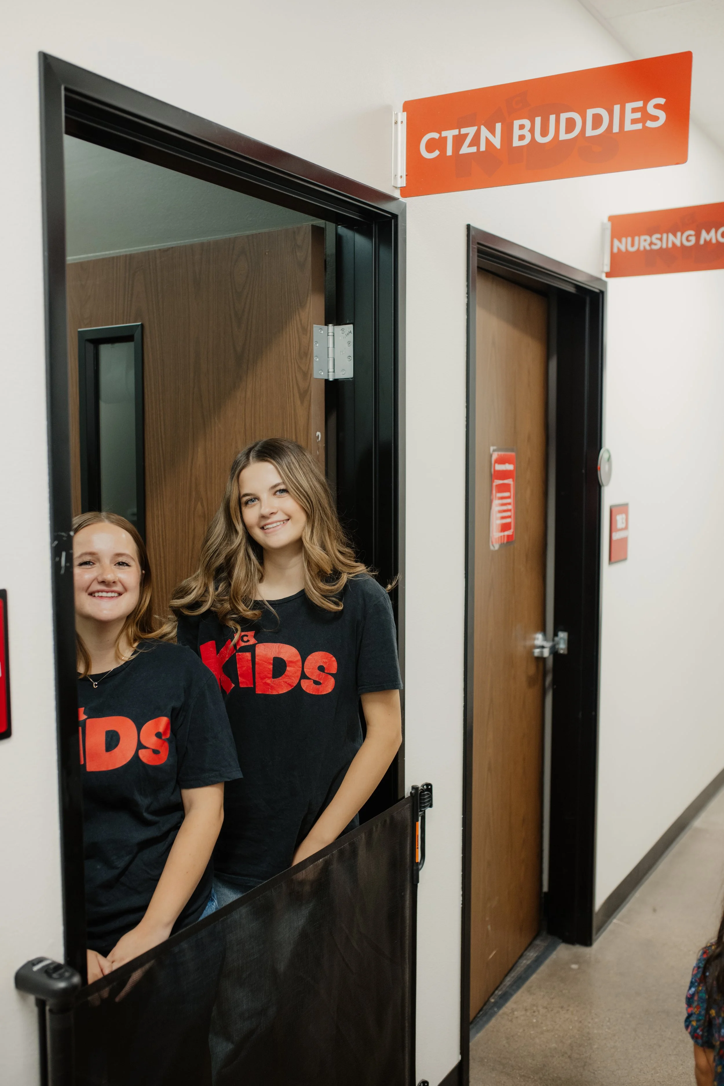 Two smiling young girls standing in front of a small partitioned area labeled 'CTZN BUDDIES' in a hallway with wooden doors and orange signs.