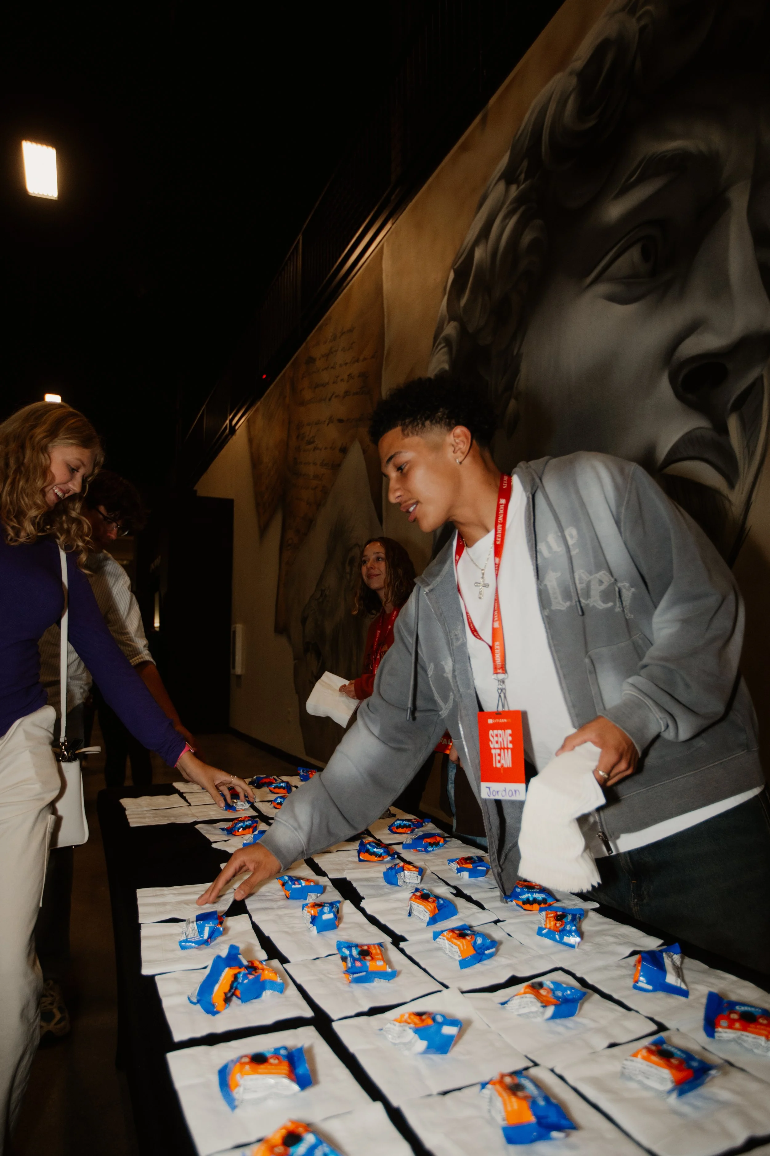People at a registration or check-in table with snacks or goodie bags, engaging in an event inside a building with a large mural of a face on the wall.