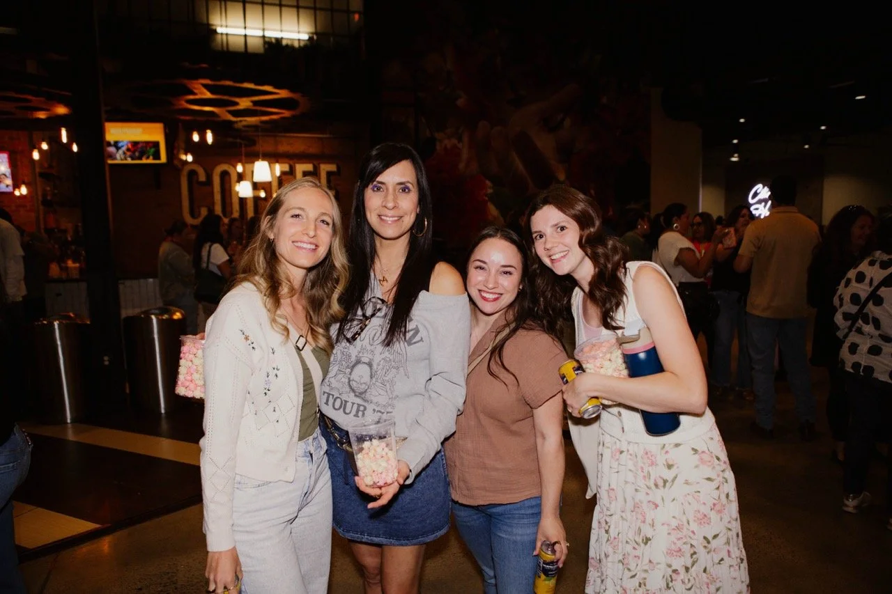 Four women smiling and holding popcorn and drinks in a crowded indoor space.
