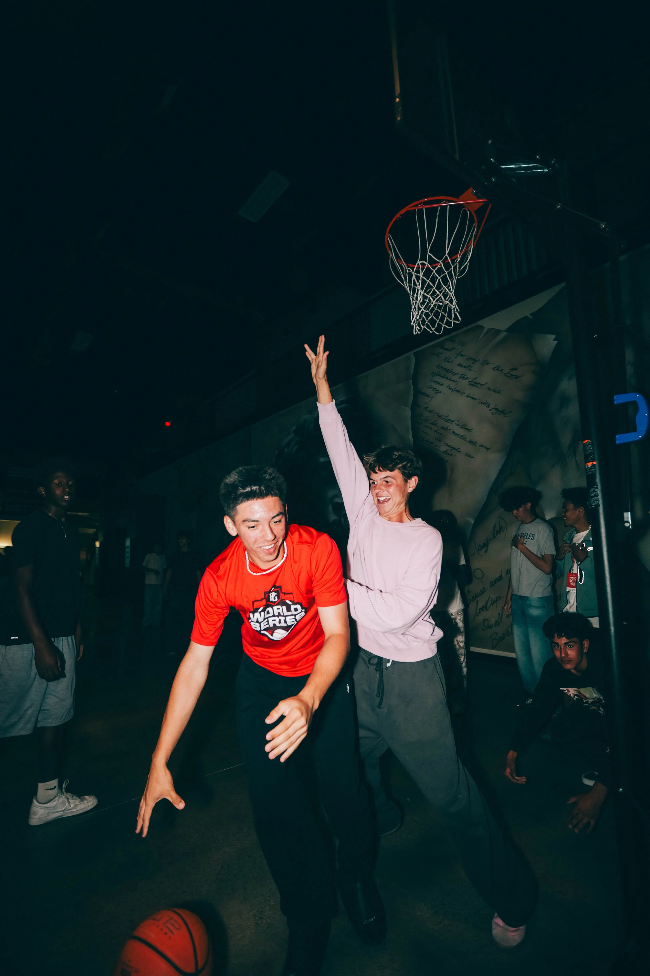 Two young people playing basketball indoors, one in a red shirt and the other in a light pink sweatshirt, with a basketball on the floor.