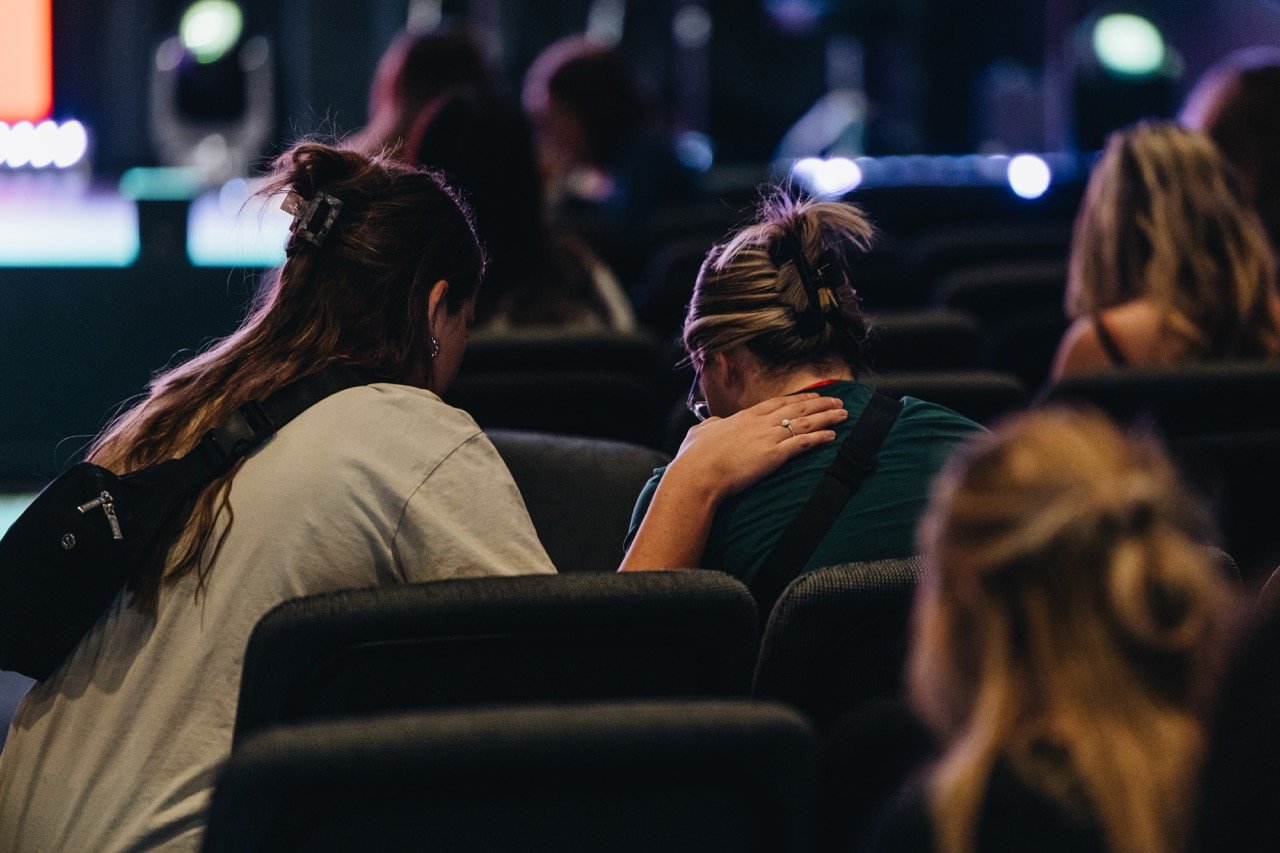 People sitting in a darkened auditorium, with two women in the foreground leaning towards each other, one whispering to the other.