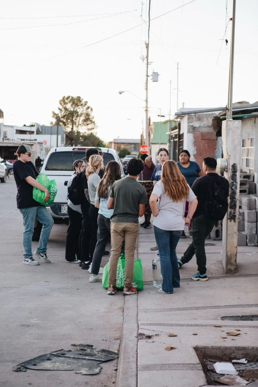Group of people standing in a line on a sidewalk in an urban area, holding bags and water bottles, with parked cars and buildings in the background.