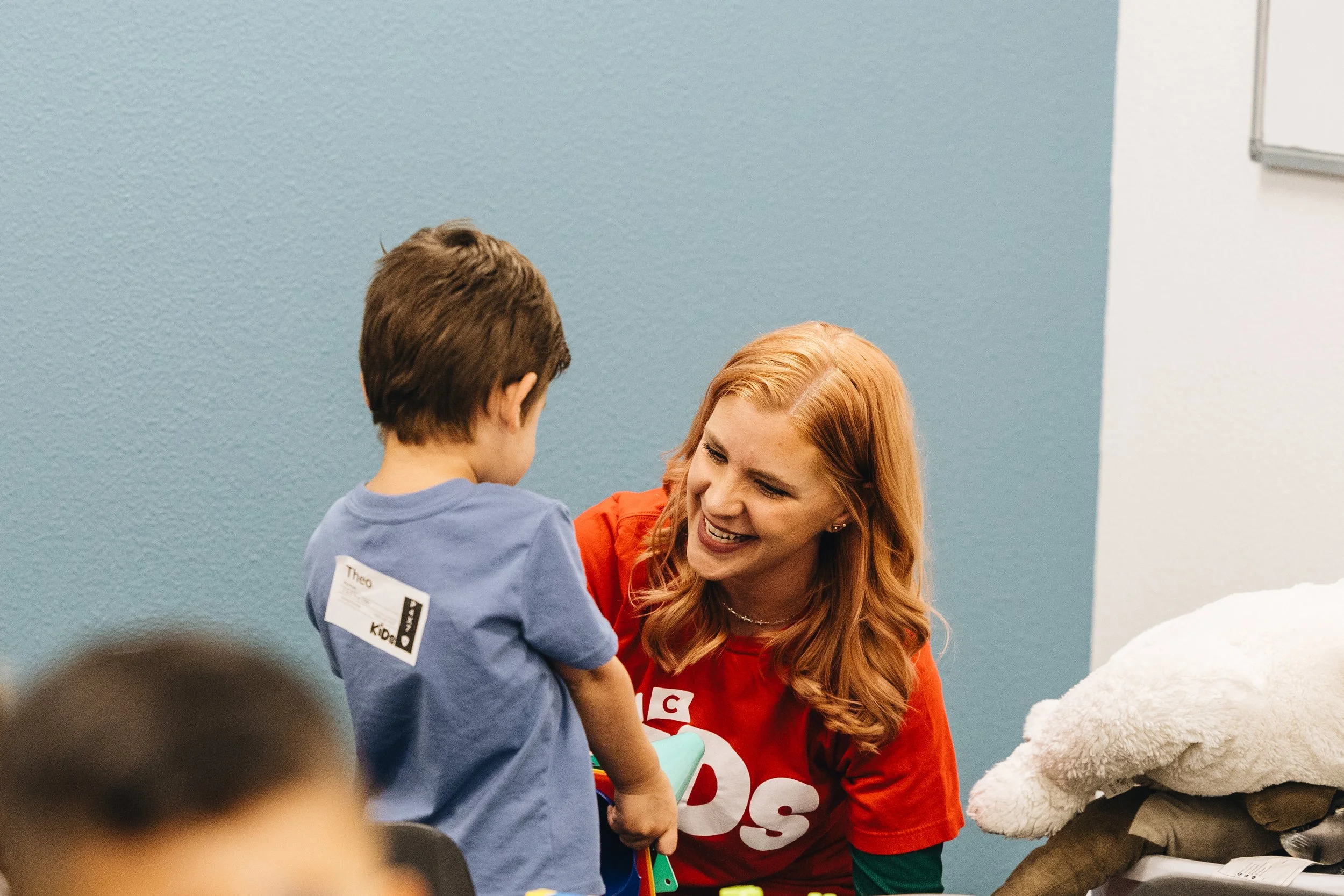 A woman with red hair smiling and talking to a young boy with a name tag on his shirt, in a classroom or community setting.